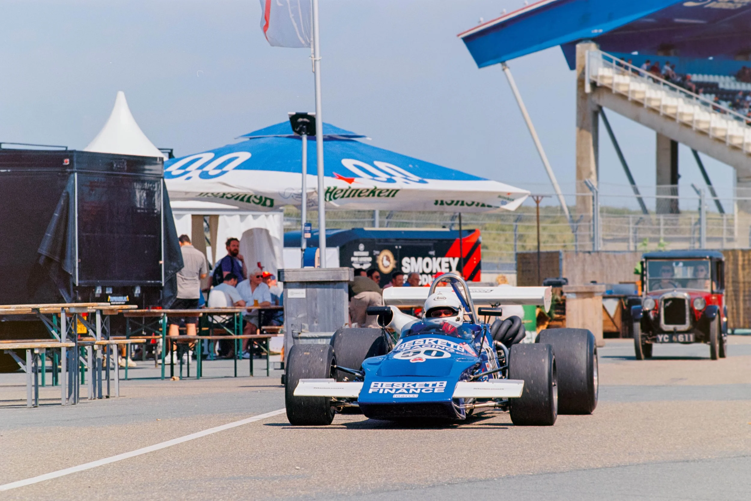 A blue race car with sponsorship logos on the front, parked in a pit area at a racetrack, with tents, a crowd, and a grandstand in the background.
