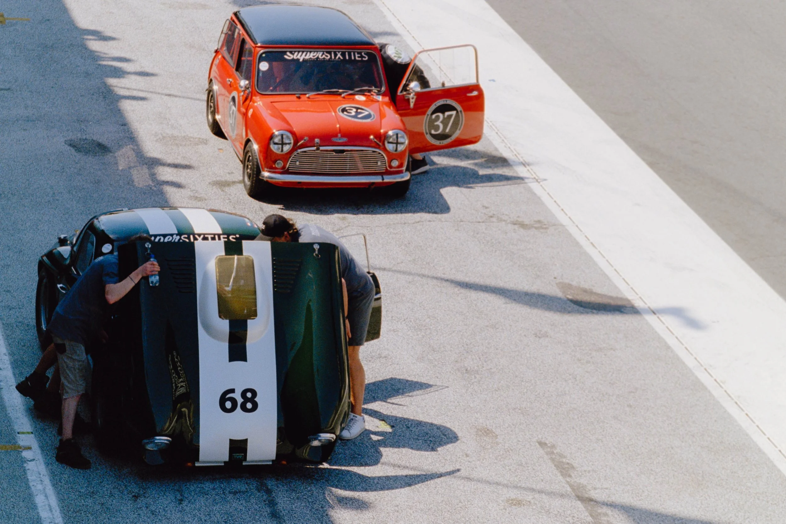 A race car is in the pit on a race track, with two crew members working on it. The car has the number 68 and a white and green color scheme, while another red race car with the number 37 is nearby with its door open.