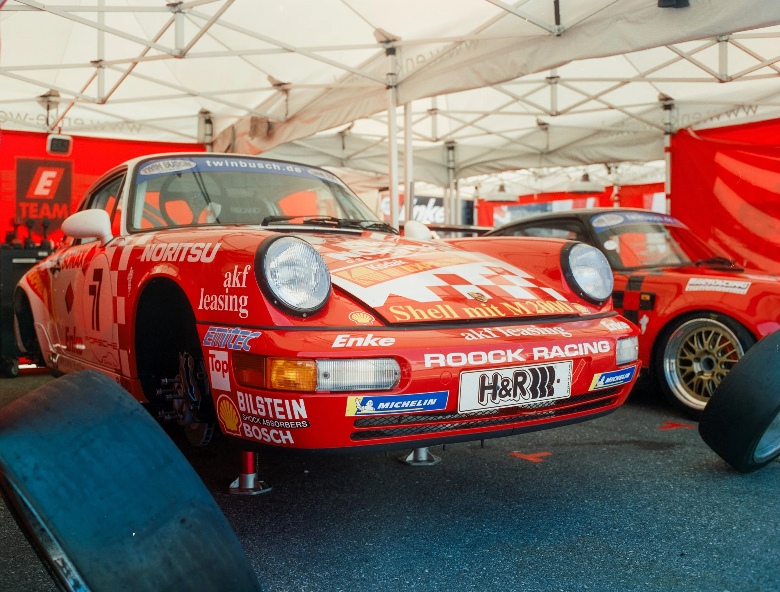Red race car under a white canopy at a motorsport event, displaying various sponsor logos and racing decals.