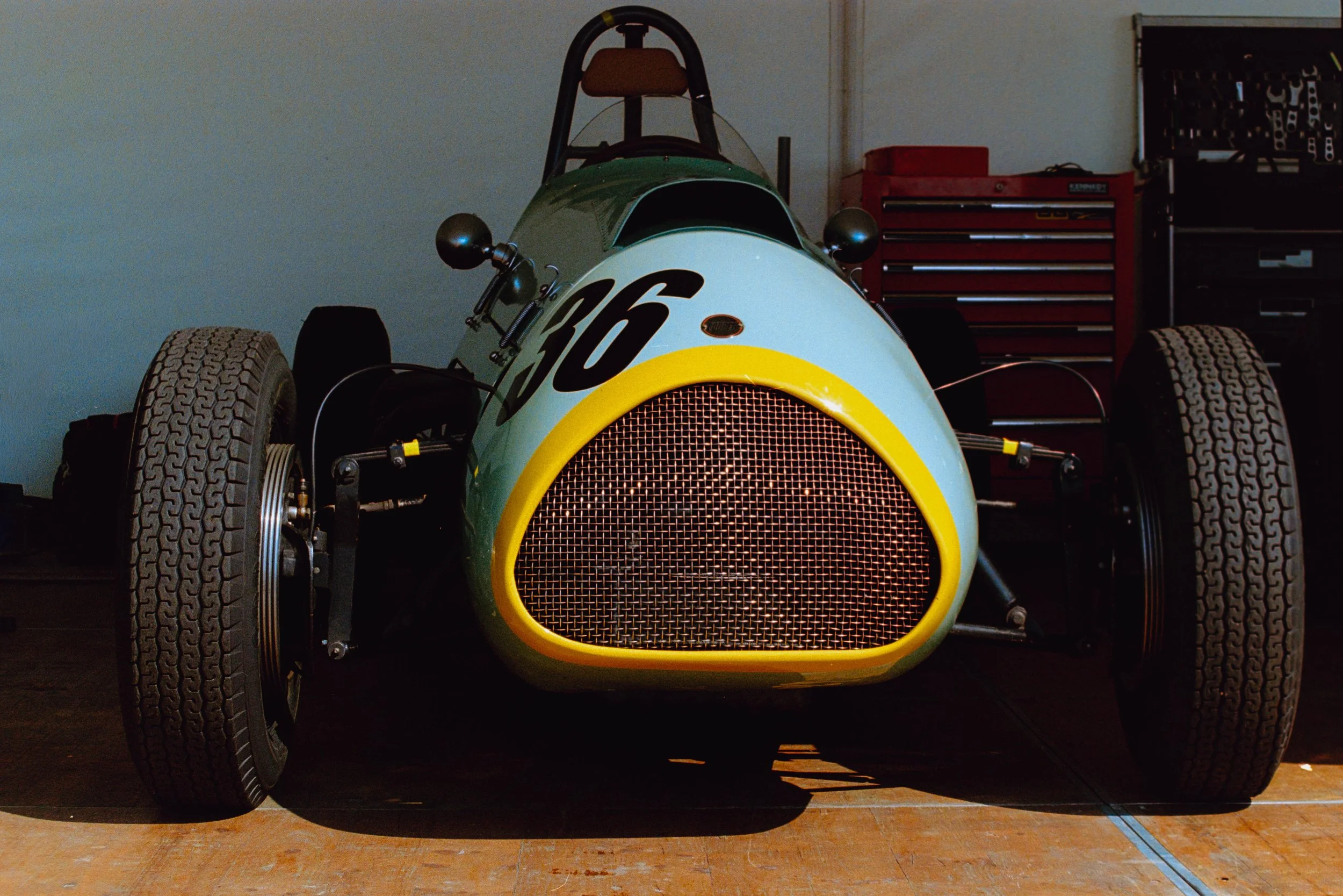 Front view of a vintage racing car with a yellow grille and the number 66 on the front, inside a workshop or garage with red tool chest and black equipment in the background.