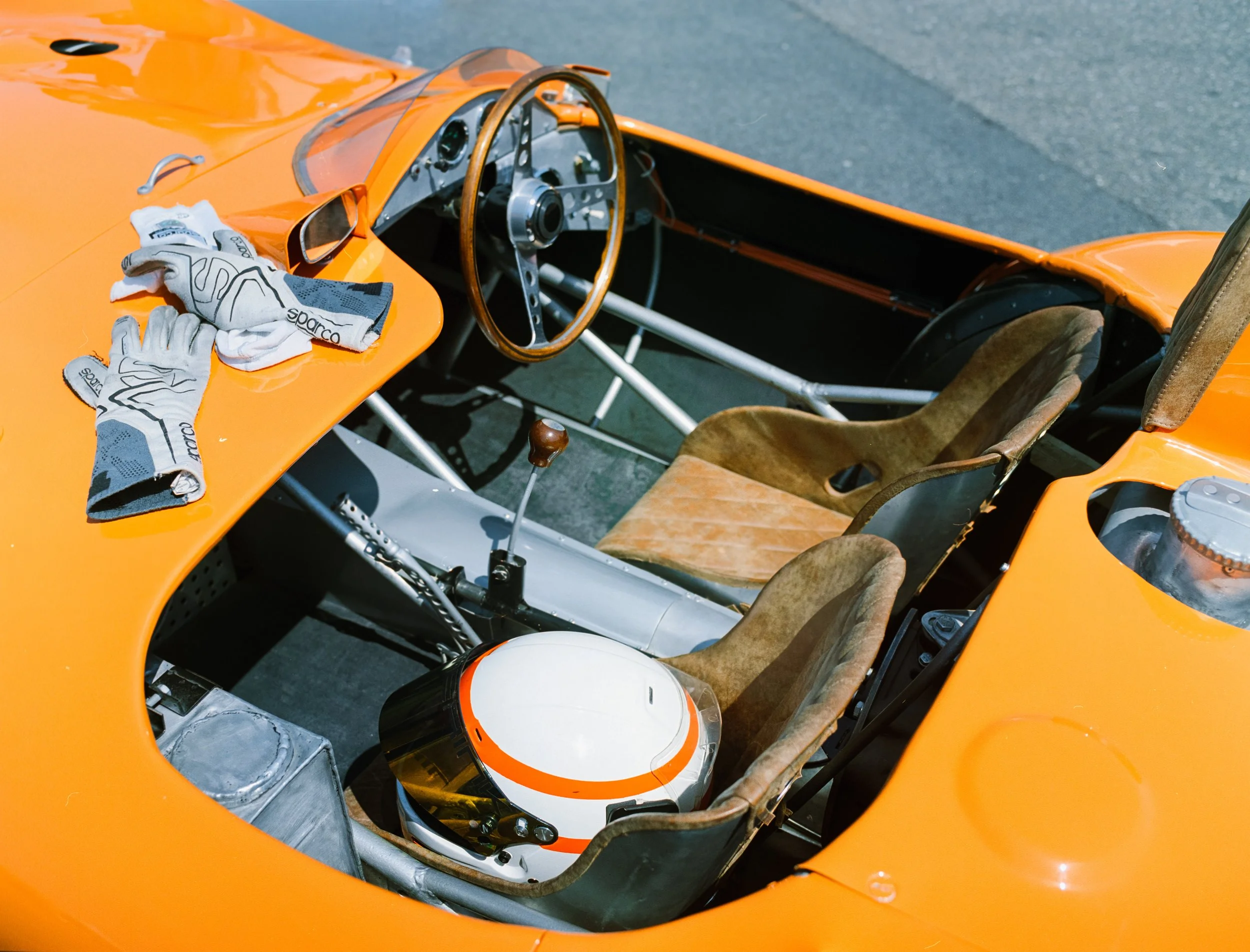 The interior of a vintage orange race car with a racing helmet on the dashboard and racing gloves on the seat.