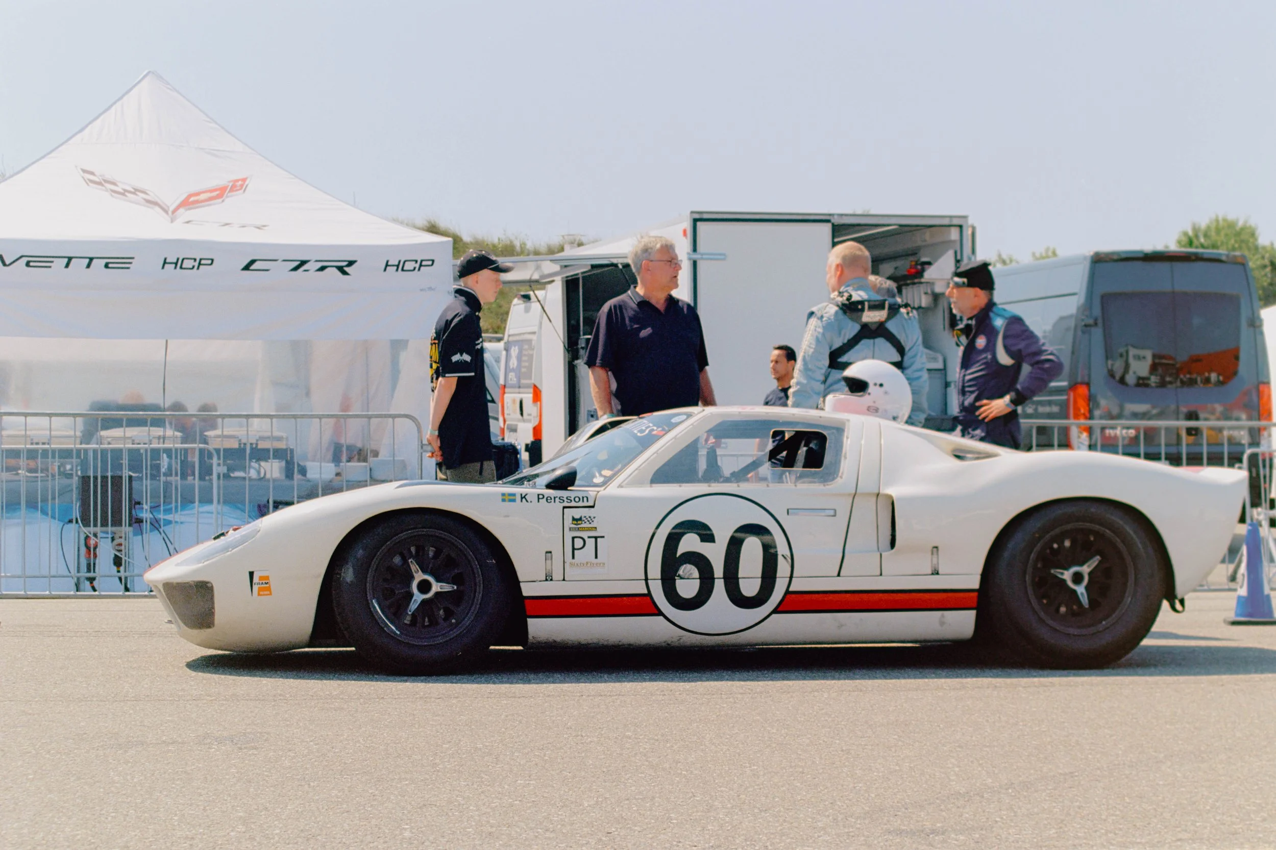 A white race car with the number 60 and the name K. Persson on the side, parked on a race track. Several people are standing nearby, including two men in racing suits and another person in casual clothing, with a tent and trailer in the background.