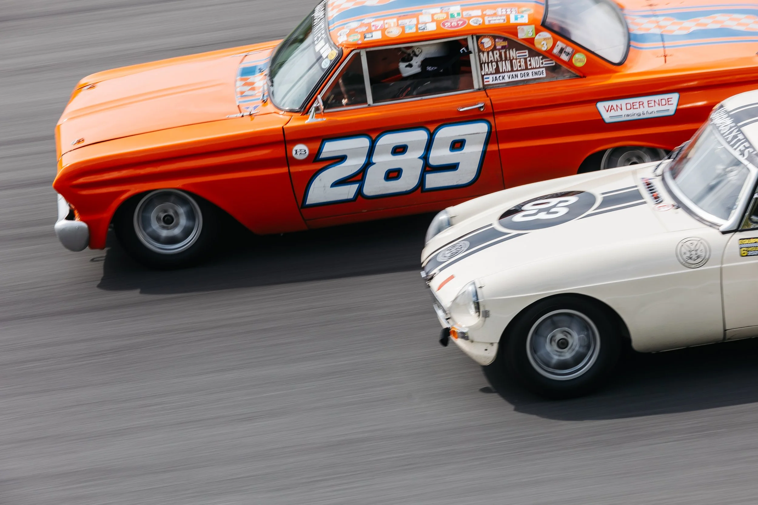 Two vintage race cars, one orange with the number 289 and another white with the number 25, speeding on a race track.