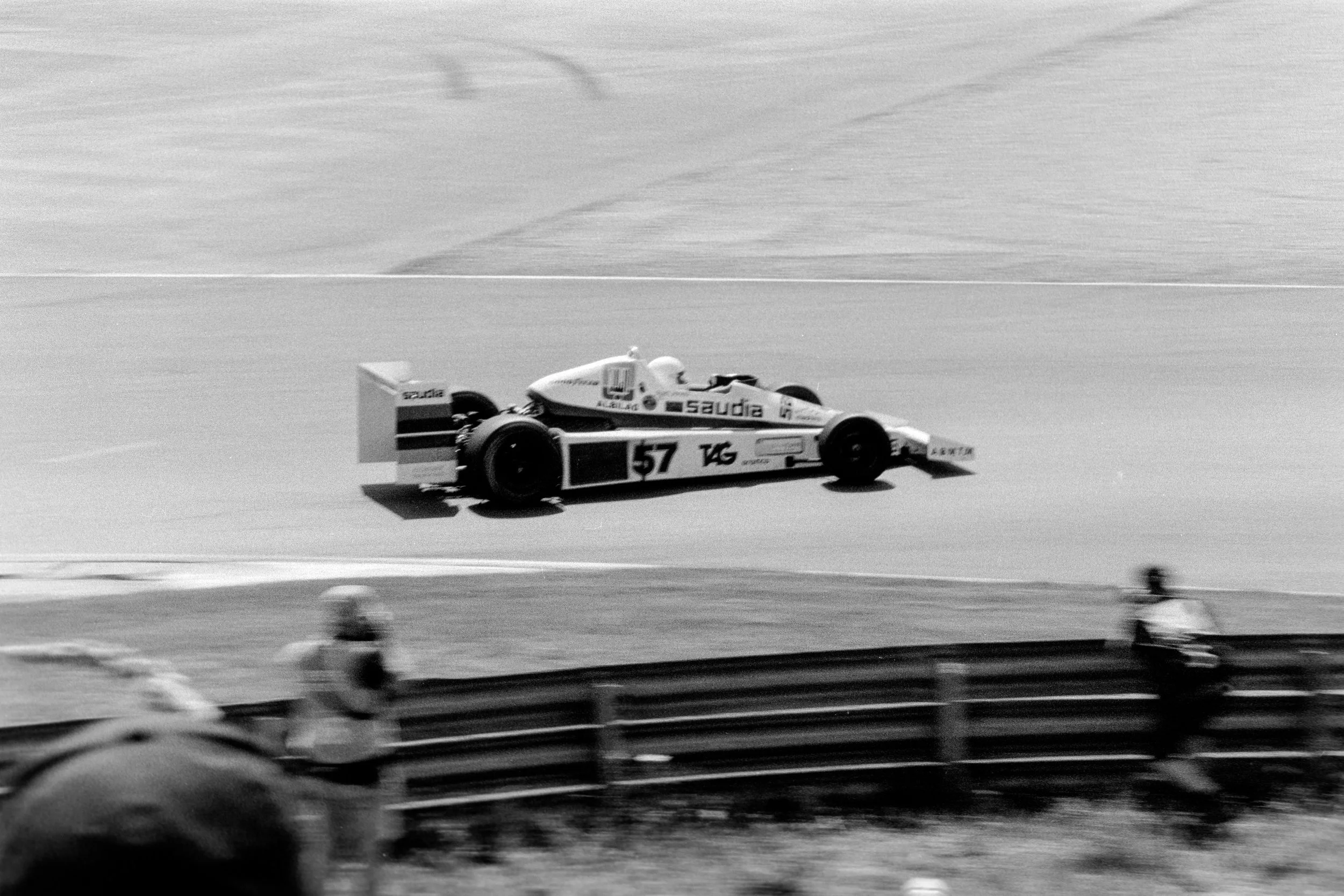 A vintage race car, number 57, on a racetrack with a flat, barren landscape in the background.