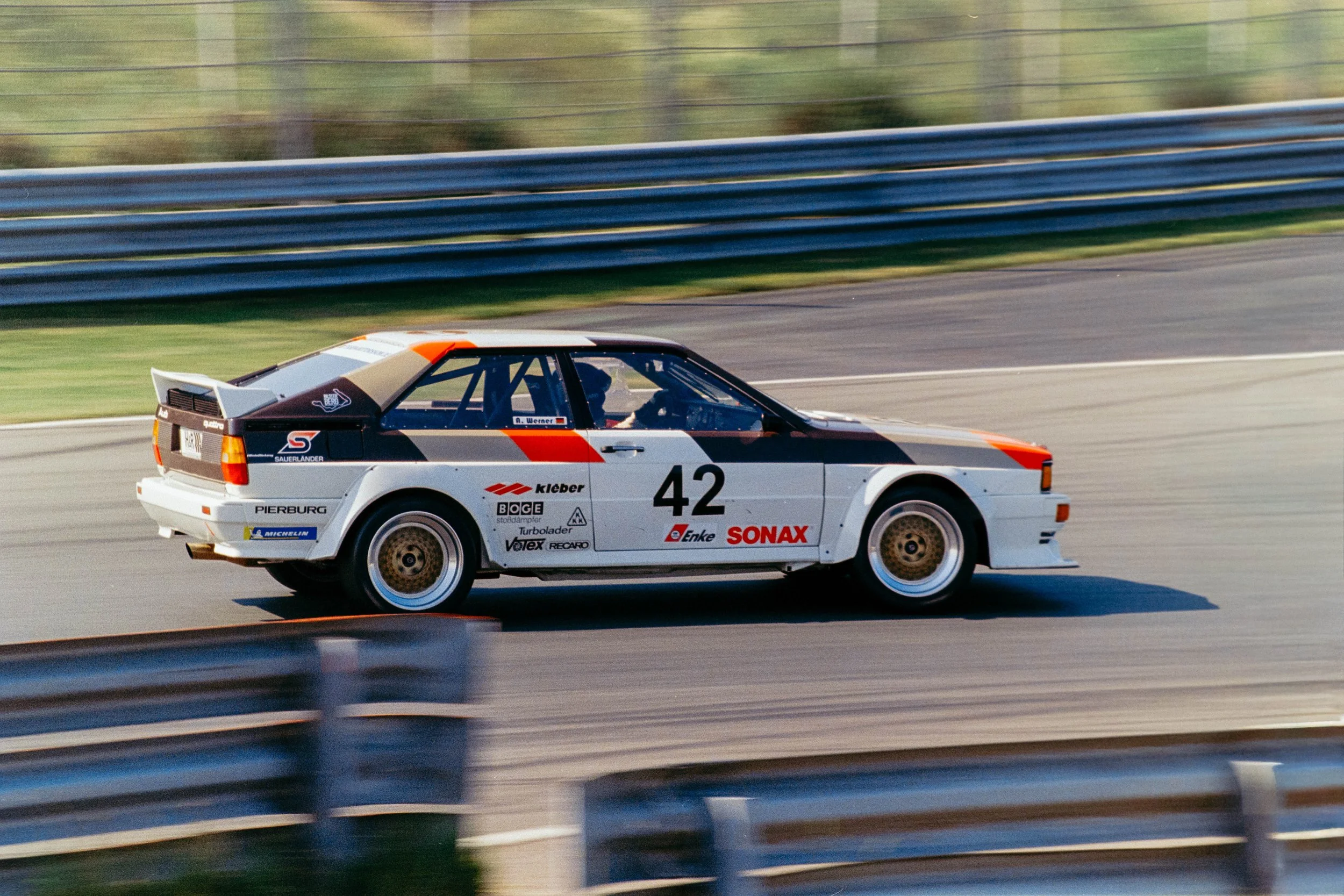 A white race car with the number 42 and various sponsor logos, driving on a race track, with safety barriers and greenery in the background.