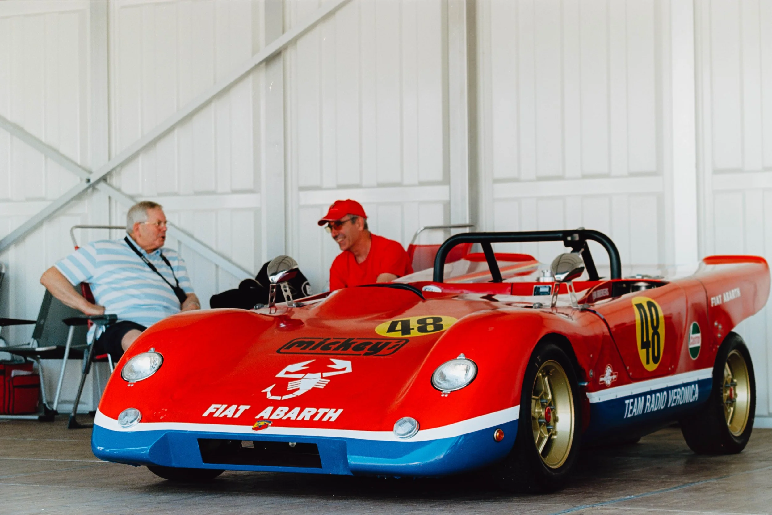 Two men seated behind a red race car inside a garage. One man has gray hair and glasses and is wearing a striped shirt, the other man is wearing a red shirt and red cap. The race car has logos including Fiat Abarth and features the number 48.