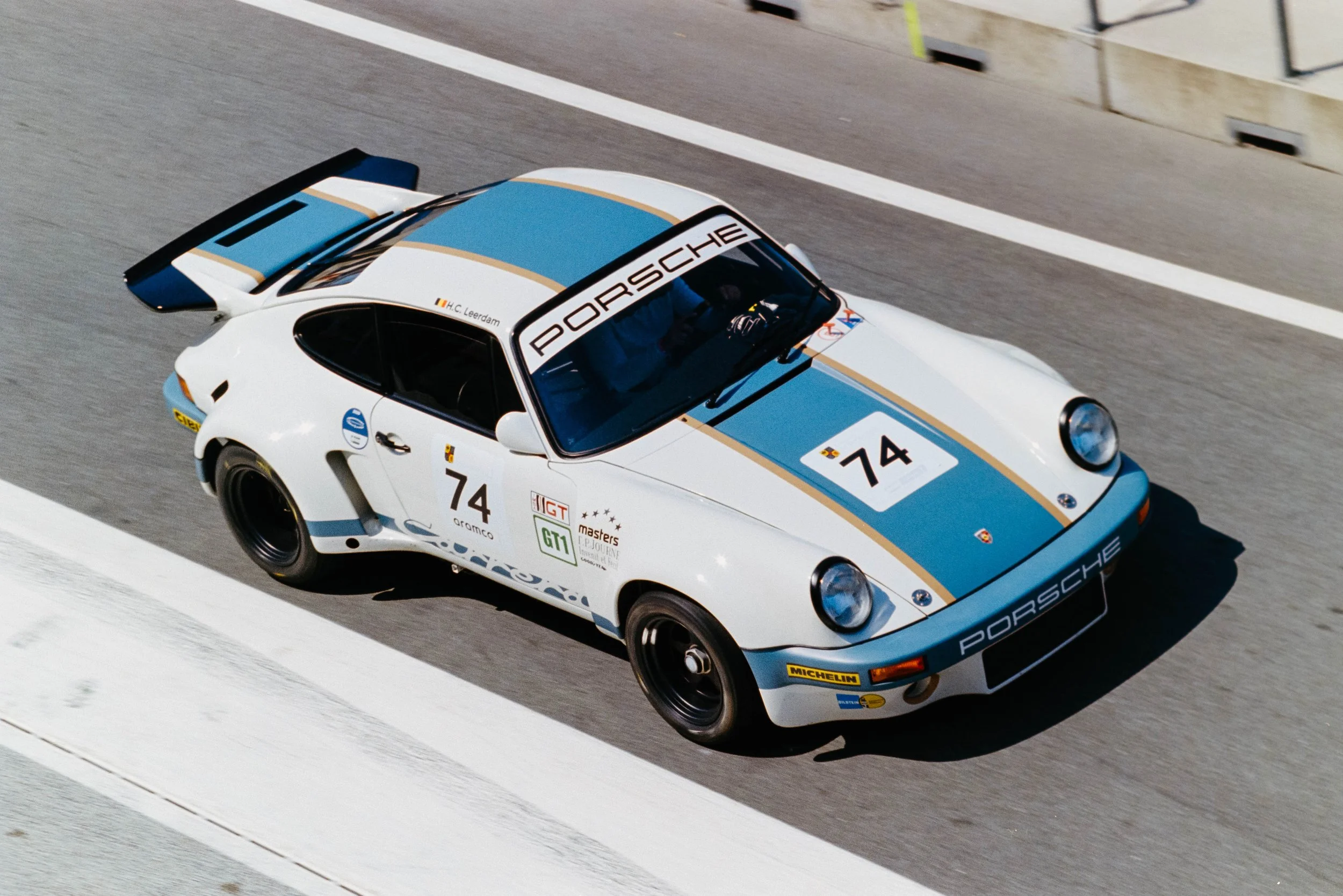 A white Porsche race car with blue and gold racing stripes, number 74, speeding on a race track.
