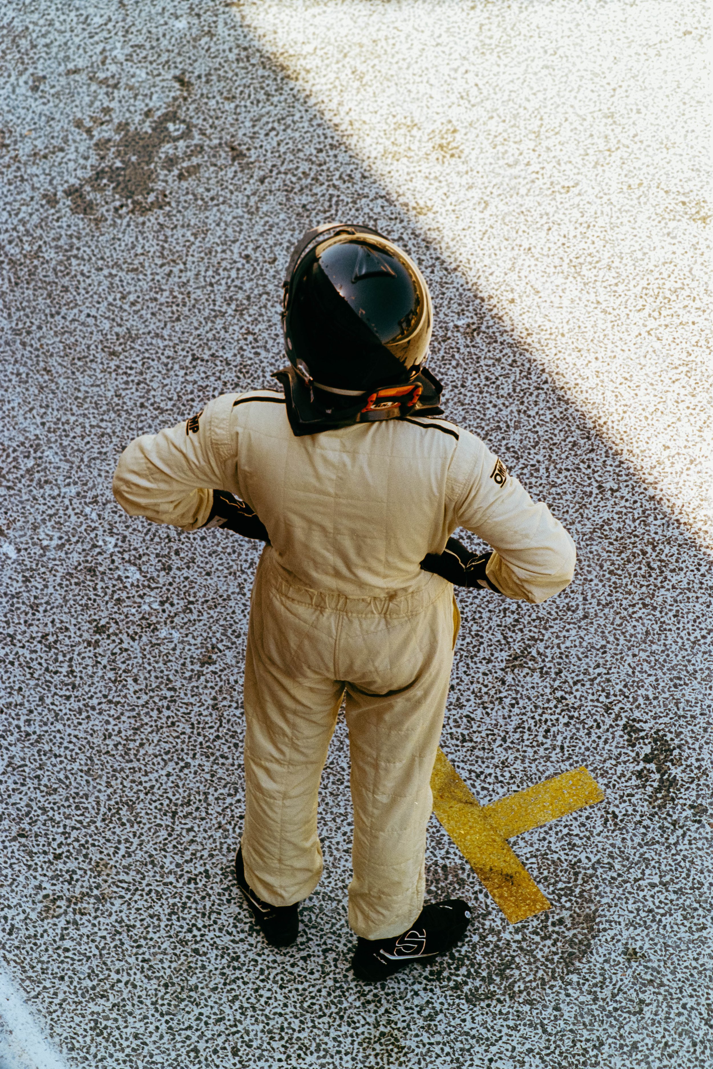 Overhead view of a person in a racing suit and helmet standing on a speckled pavement next to a yellow line in a parking lot.