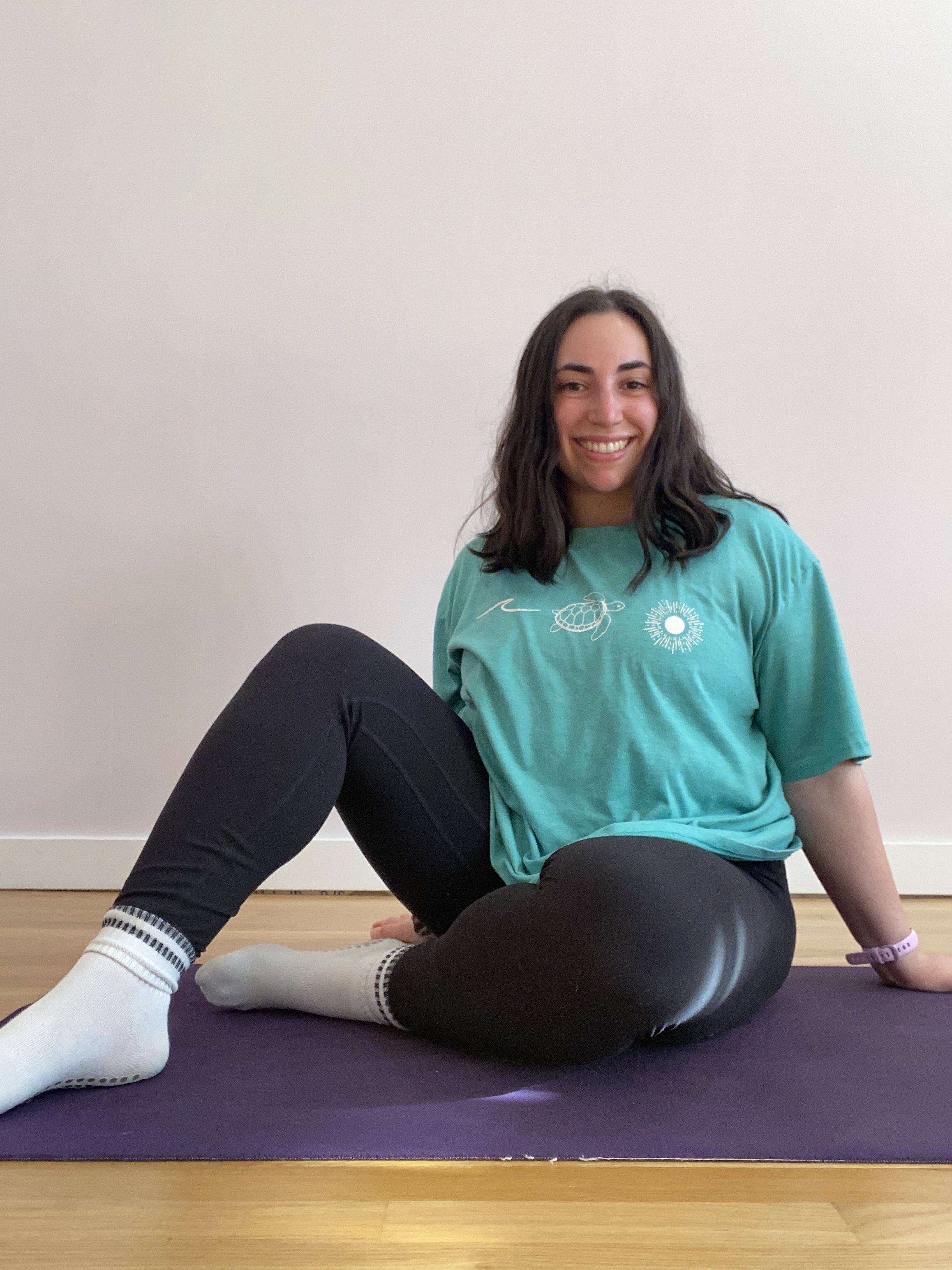 Young woman with dark hair sitting on Pilates mat,  posing with right leg up, smiling, wearing a teal T-shirt and black leggings, against a plain white wall.