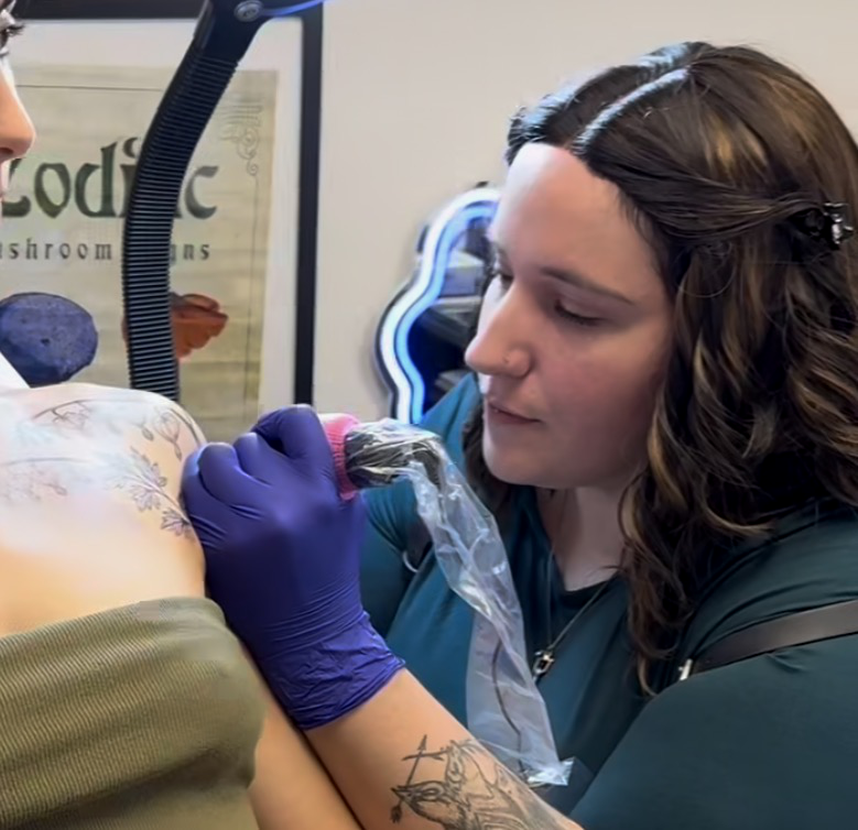 A tattoo artist wearing purple gloves working on a woman's shoulder tattoo in a tattoo studio.