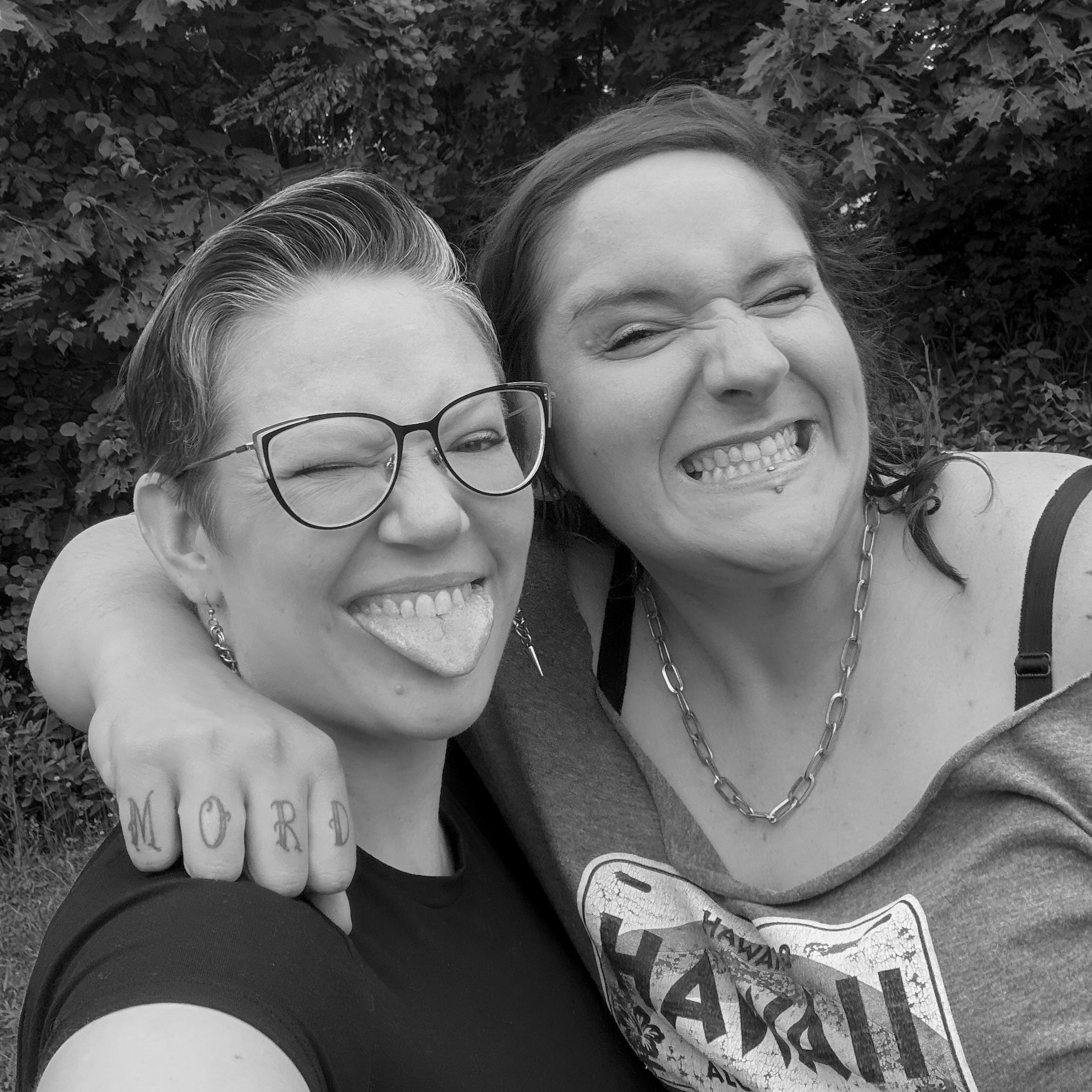Close-up of two smiling women, one wearing glasses and a tank top, and the other with long hair and a chain necklace, outdoors with trees in the background.