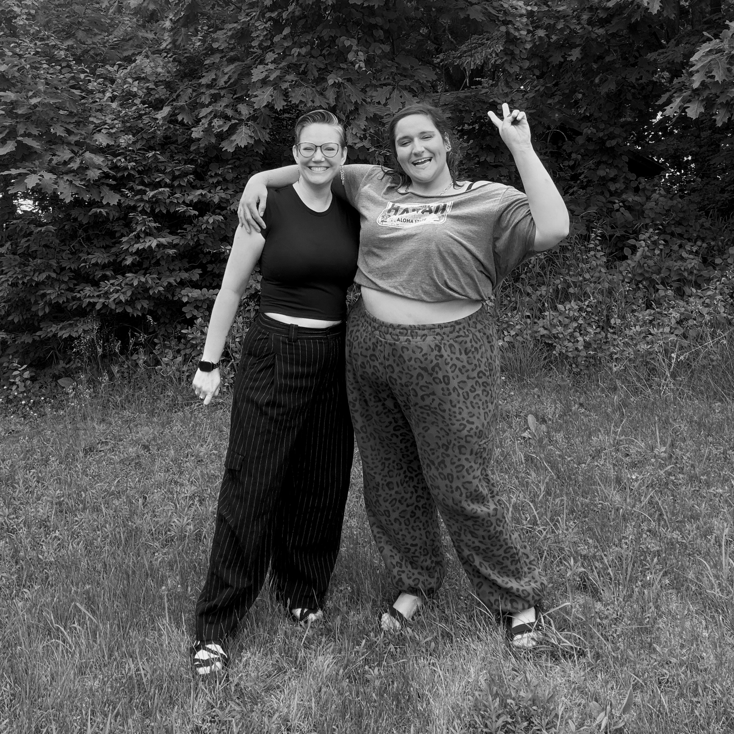 Two women standing outdoors in front of dense foliage, with arms around each other, smiling and making peace signs, in black and white.