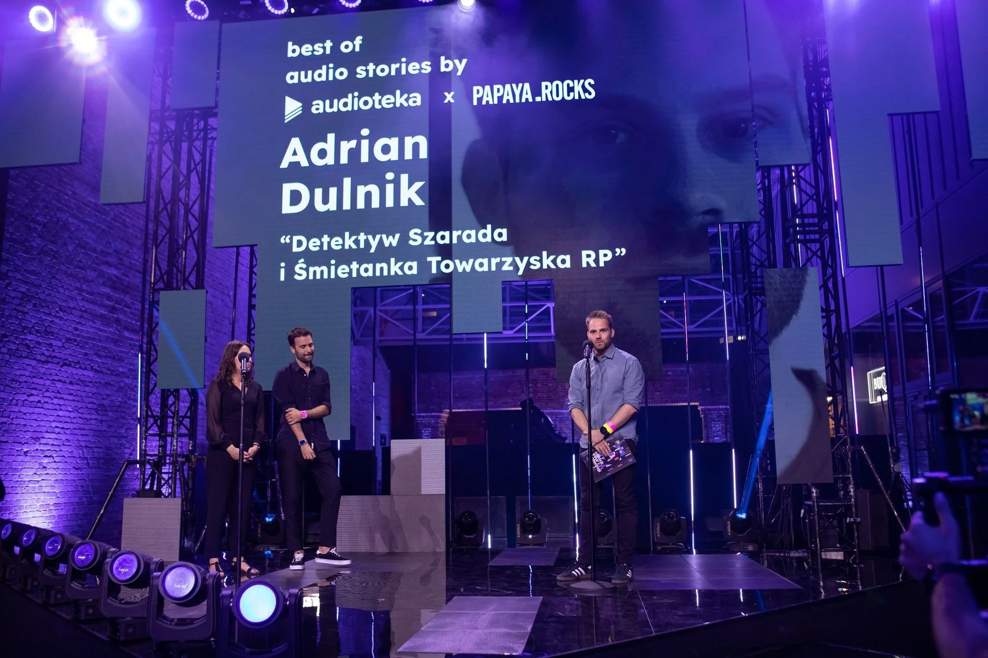 Group of three people standing on a stage with a large screen behind them displaying text about audio stories by audioteka and Papaya.Rocks, and the title Adrian Dulnik, with a woman and two men, one of whom is holding a book, in an event setting with purple lighting and stage equipment.