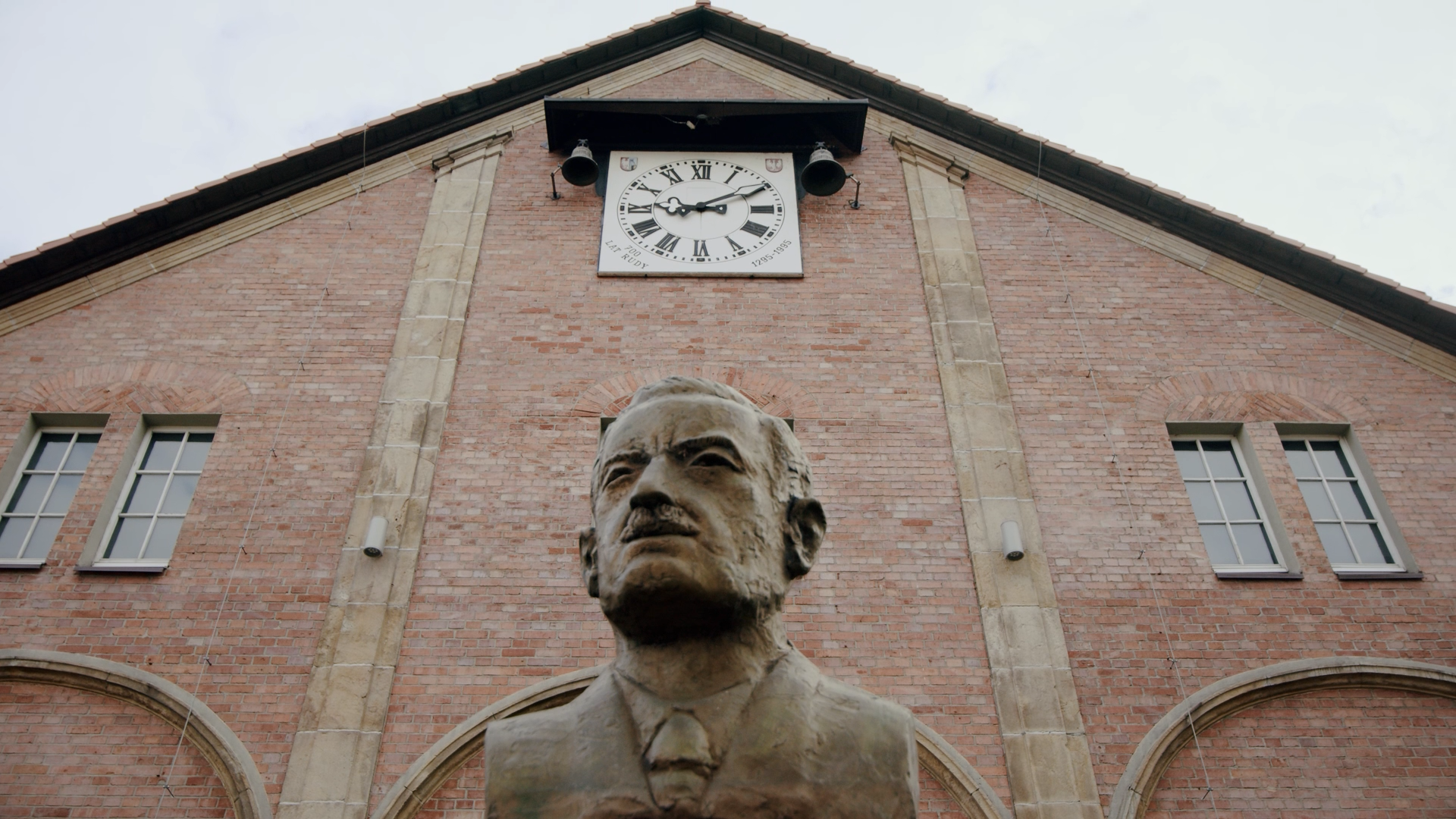 A brick building with four rectangular windows and a large clock showing the time, with a bust sculpture of a man in front of it.