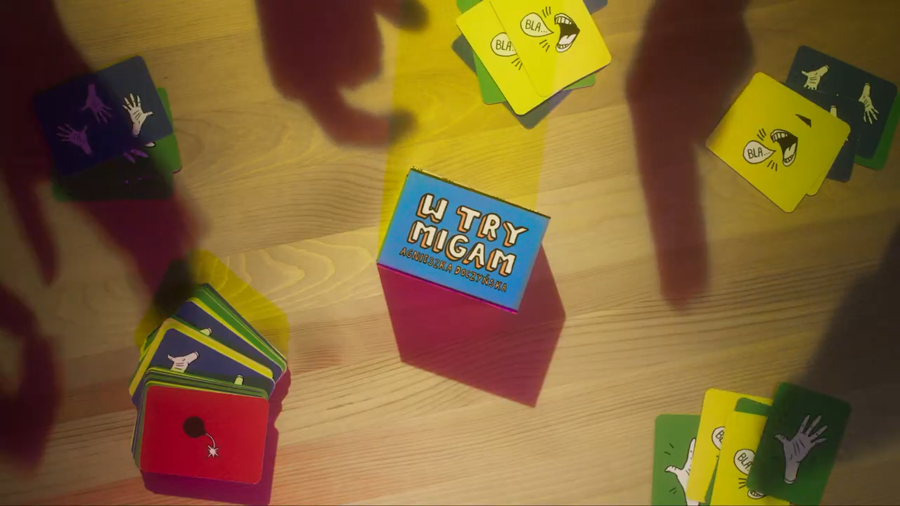 Card game called 'Try Migam' by Agnieszka Doczyńska, with colorful cards featuring illustrations of hands, speech bubbles with 'BLAH', and a red card with a bomb illustration, spread on a wooden surface.