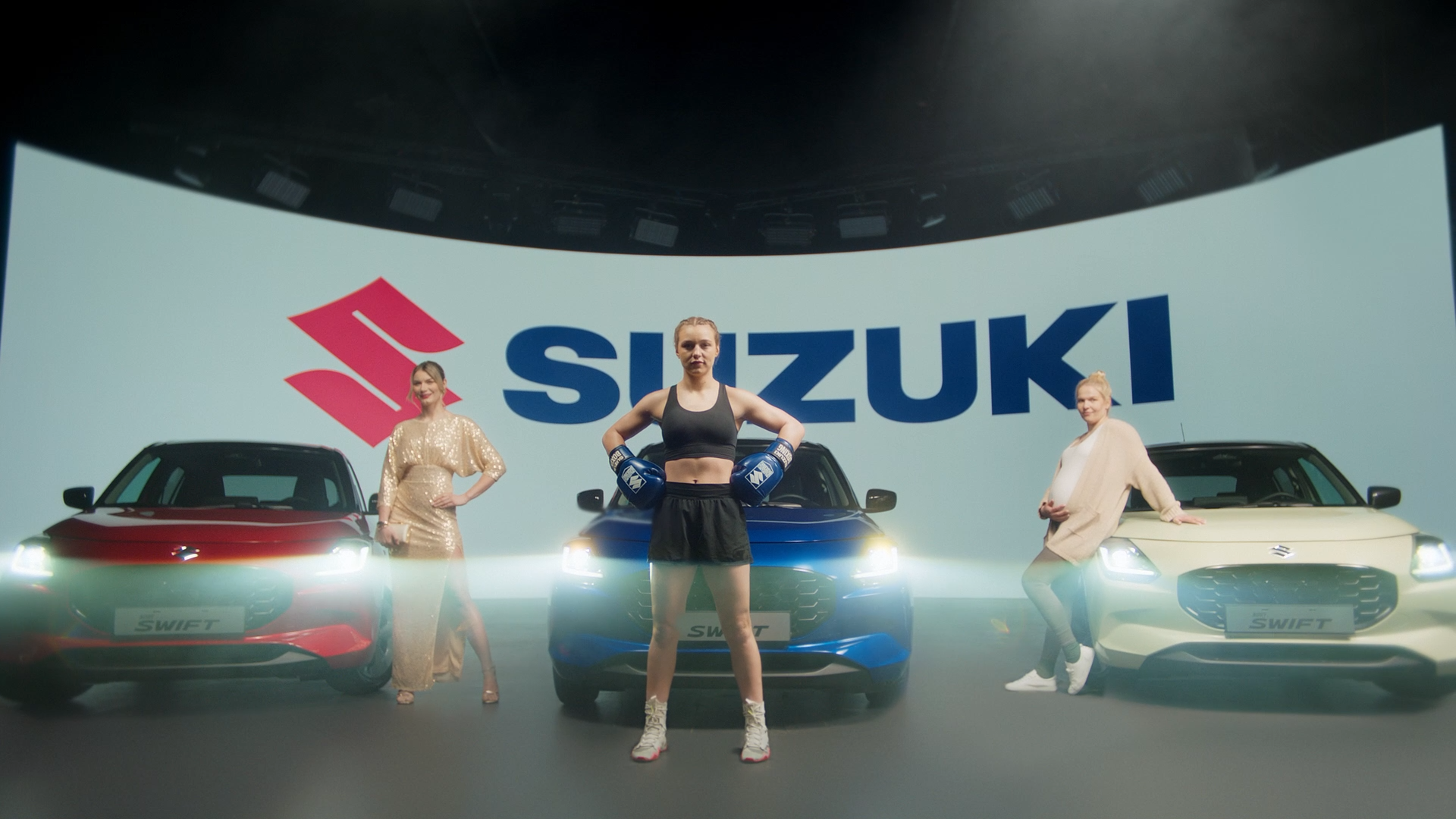 Three women and three Suzuki Swift cars on stage at a launch event. The woman in the center wears boxing gloves and sportswear, while the other two women are dressed in fashionable outfits. A large Suzuki logo is displayed in the background.