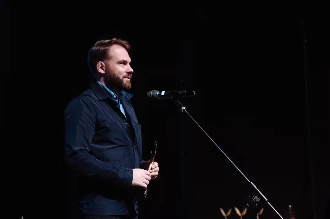 A man with a beard and brown hair stands on stage, wearing a dark jacket and holding glasses, speaking into a microphone against a black background.