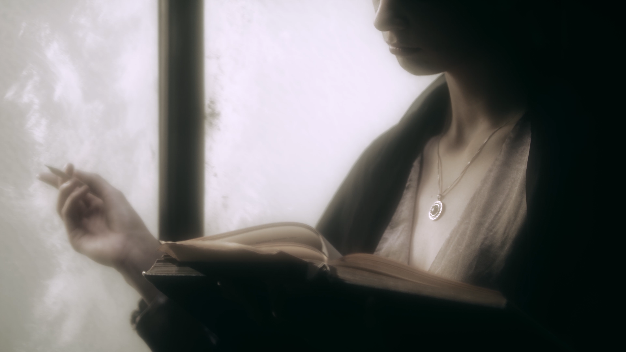 A woman is reading a book or magazine while sitting near a window, with natural light illuminating her face and upper body. She is wearing a necklace and a dress or blouse.