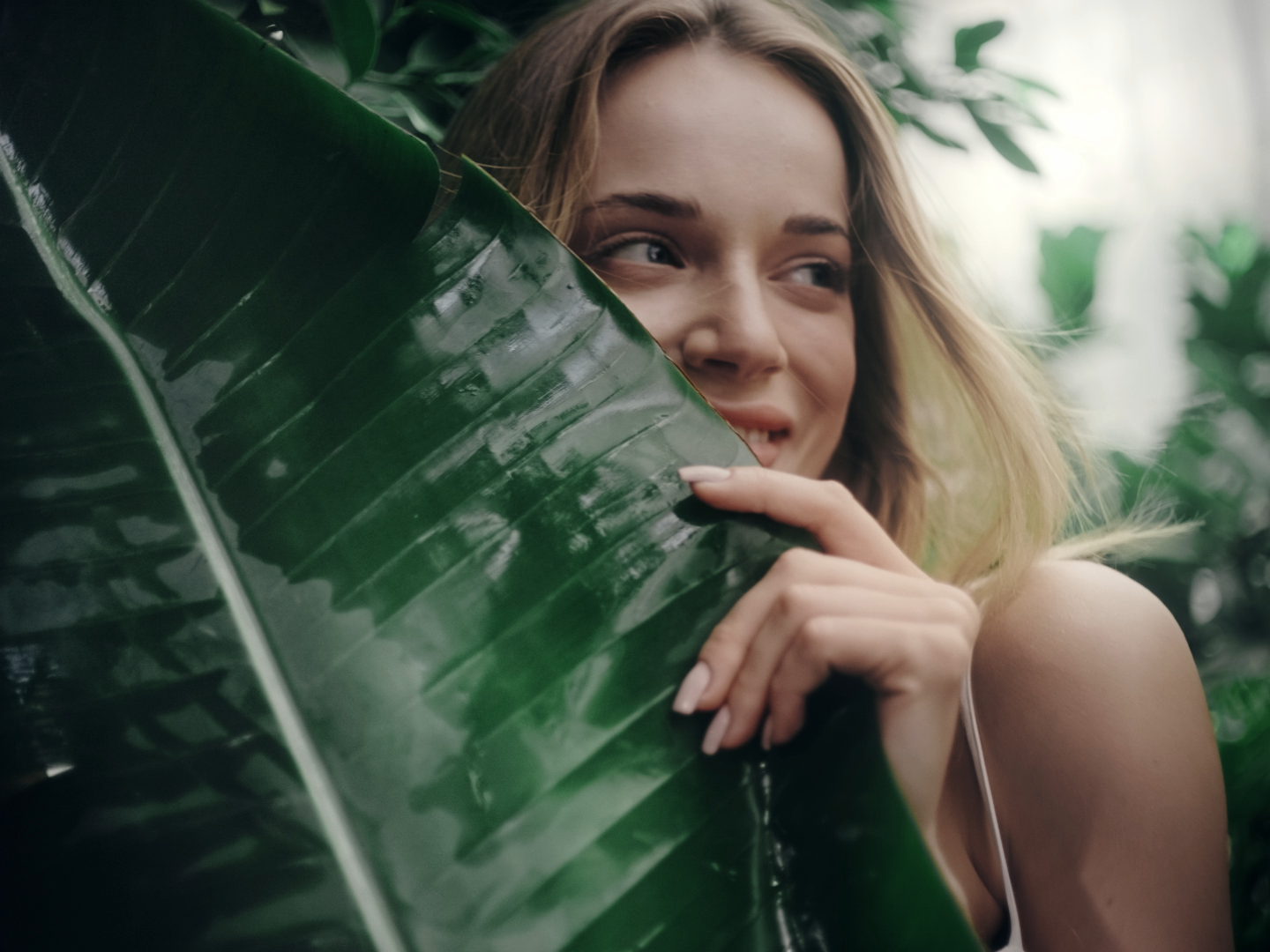 A young woman with blonde hair smiling and partially hiding behind a large green tropical leaf in a lush, green environment.
