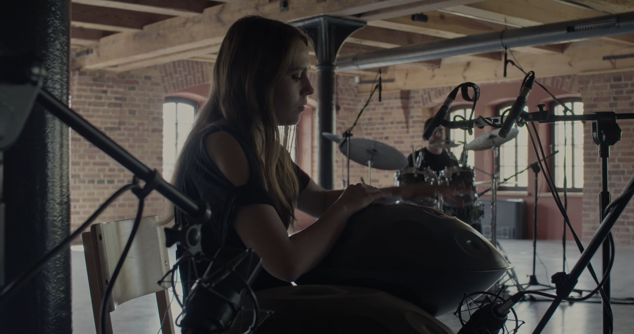 A young woman playing a steel drum in an industrial-style room with exposed brick walls and large windows. A drummer is visible in the background.