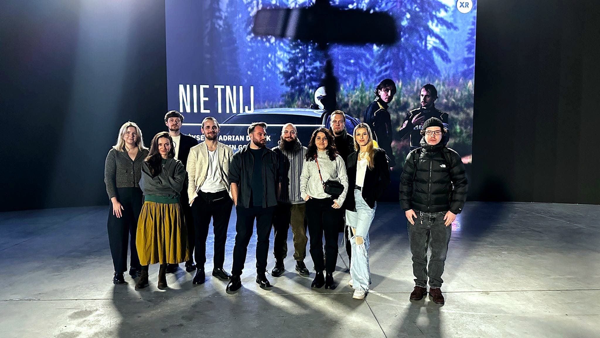 A group of ten people standing in front of a large display screen at an event or exhibition, with a forest scene and the text 'NIET NIJ' visible on the screen.