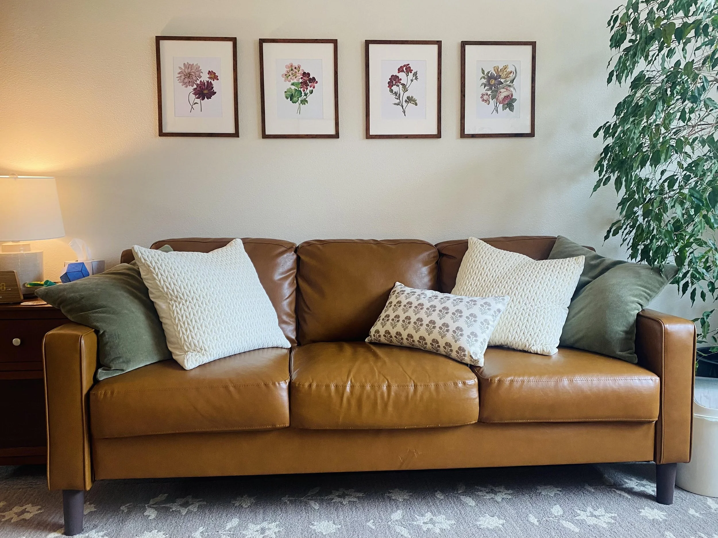 Mary Corelli's therapy office with a brown leather sofa featuring three pillows and a side table with a lamp and tissue box. Above the sofa are four framed floral prints on a light-colored wall, and to the right, a tall green plant.