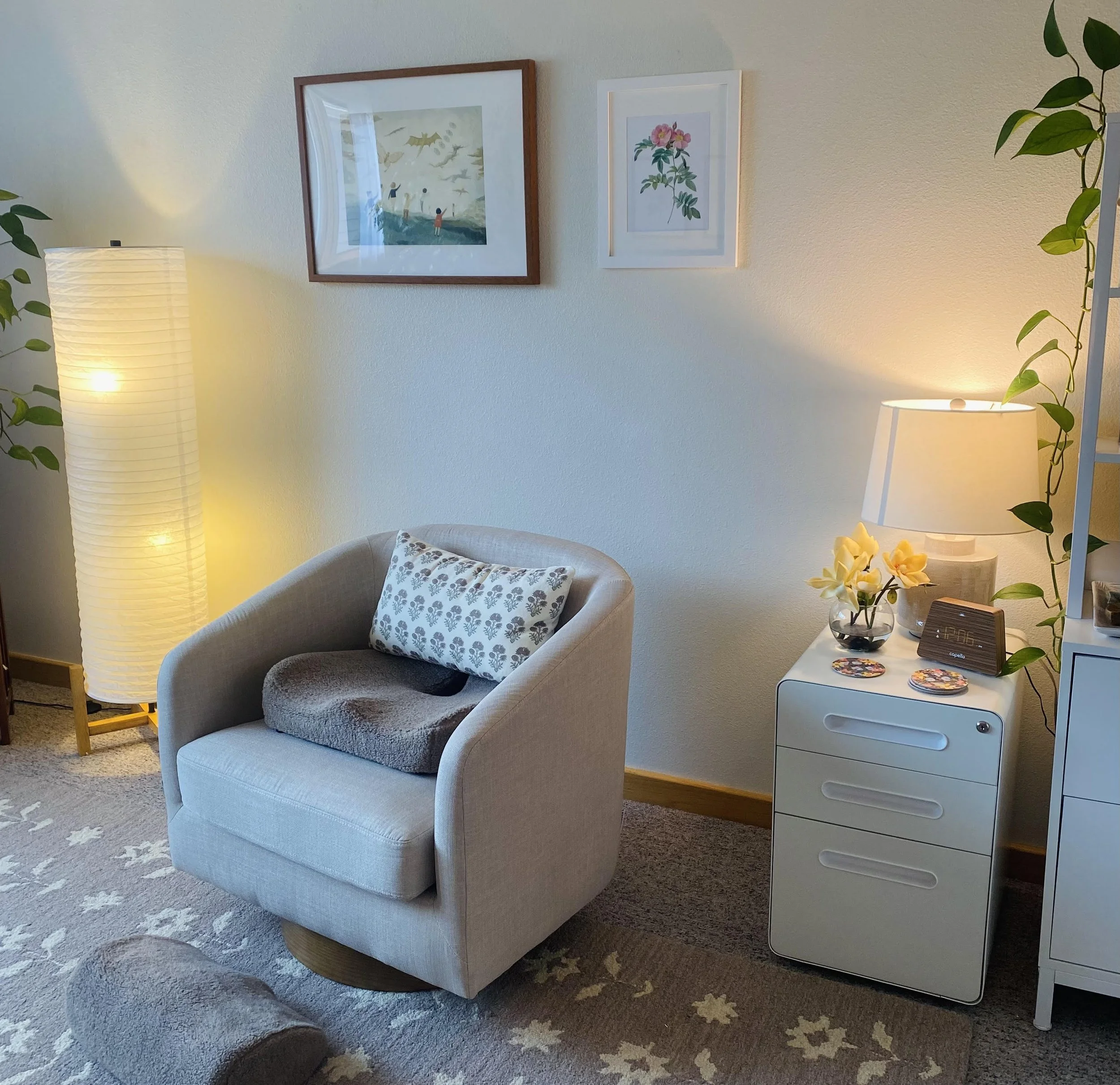 Mary Corelli's therapy office with a beige armchair and floral pillow aside a softly glowing floor lamp. On the wall above hang two paintings in muted colors. To the right, a green leafy vine hangs near a vase of cream-colored flowers and a clock.