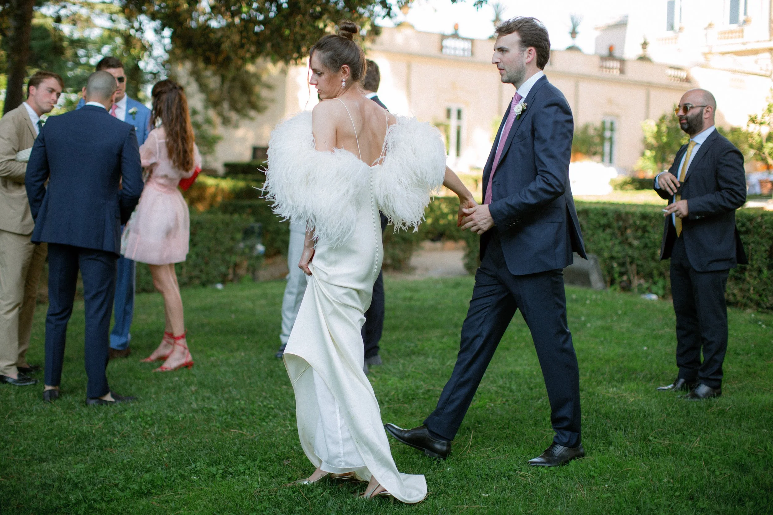 Un matrimonio elegante si tiene al centro dell'immagine, la sposa indossa un abito bianco lungo e una pelliccia bianca mentre il marito indossa un completo blu scuro con cravatta rosa. Sullo sfondo, altri invitati sono riuniti su un prato verde, con 