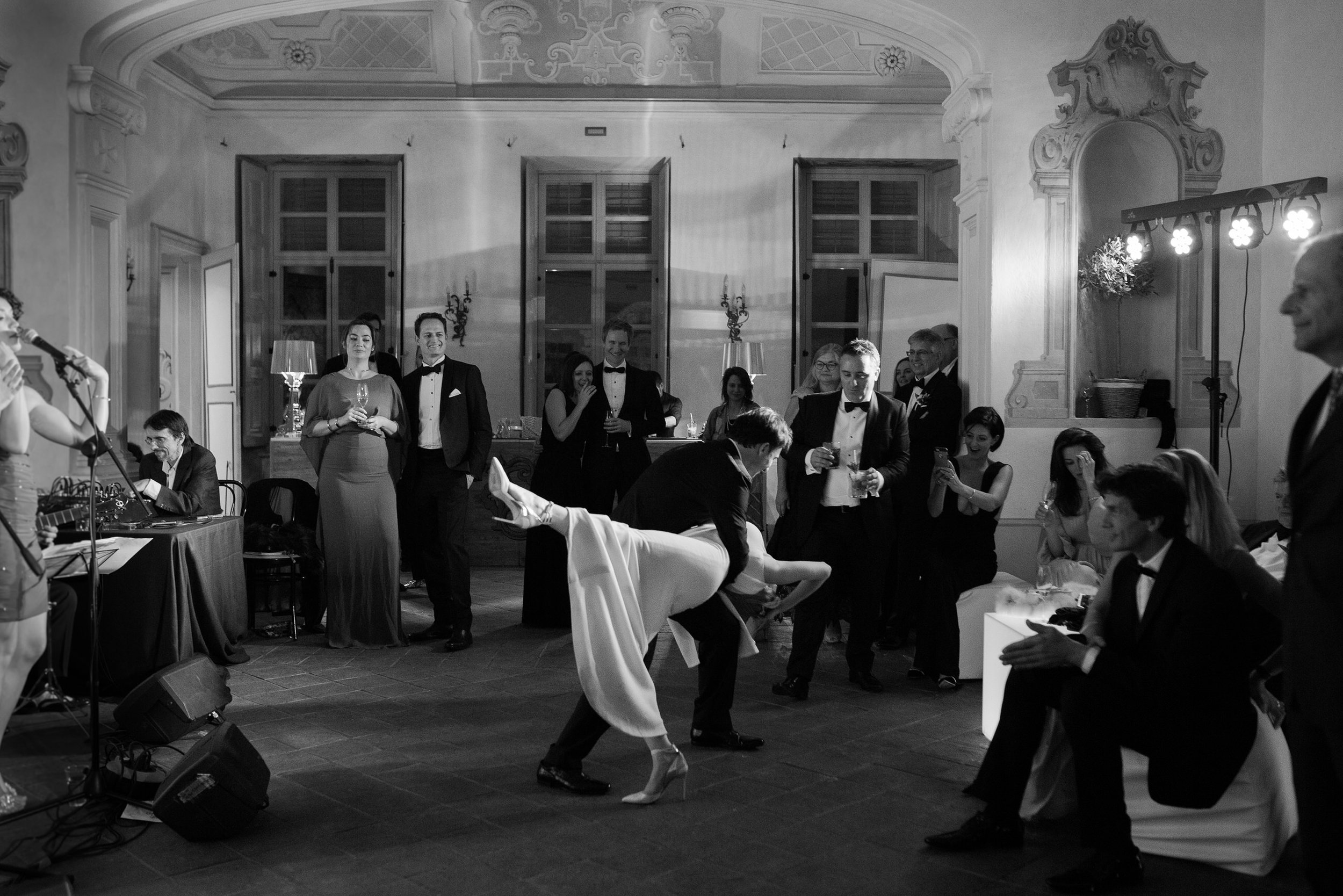 Una scena di un ricevimento elegante con persone vestite in abiti formali, alcuni che brindano e altri che ballano in una sala decorata con elementi classici e luci calde.