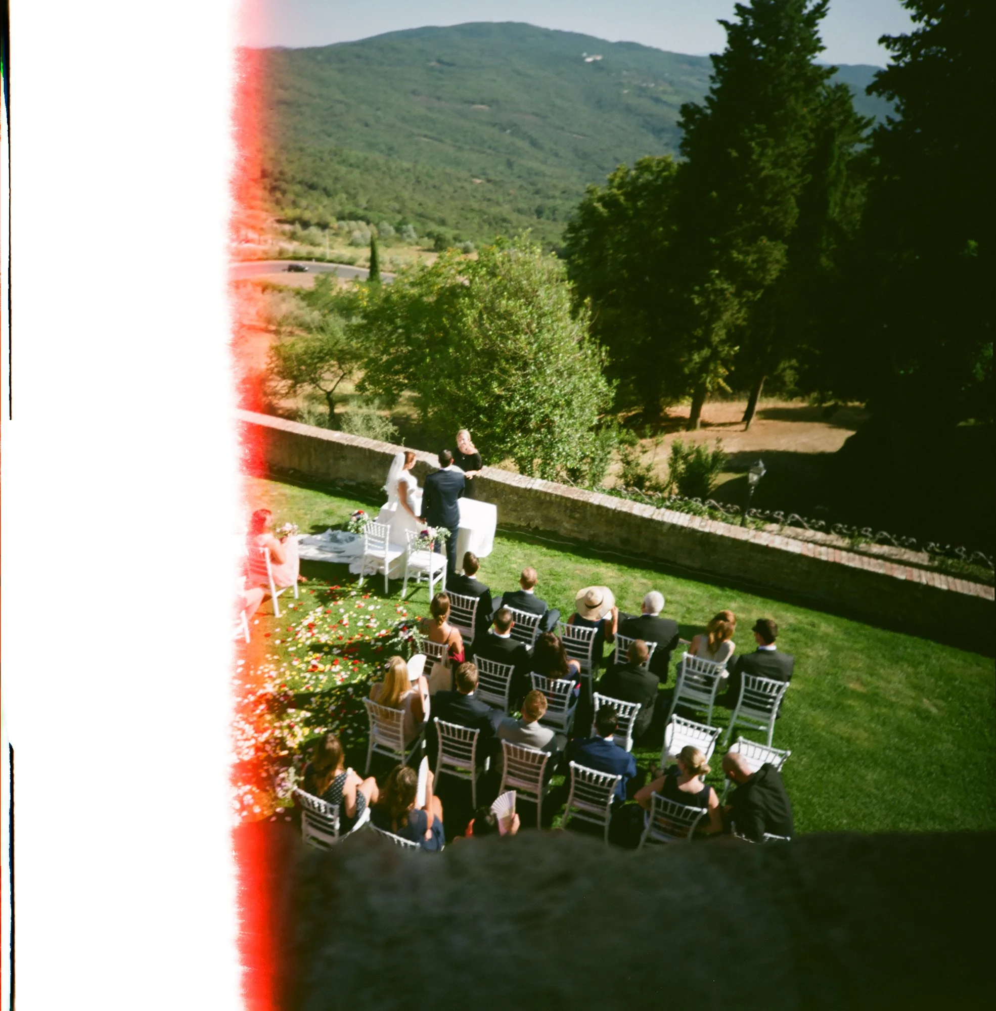 Un matrimonio che si sposa all'aperto su un prato con alle spalle delle montagne verdi, gli invitati seduti in sedie bianche, decorazioni floreali e un cielo soleggiato.