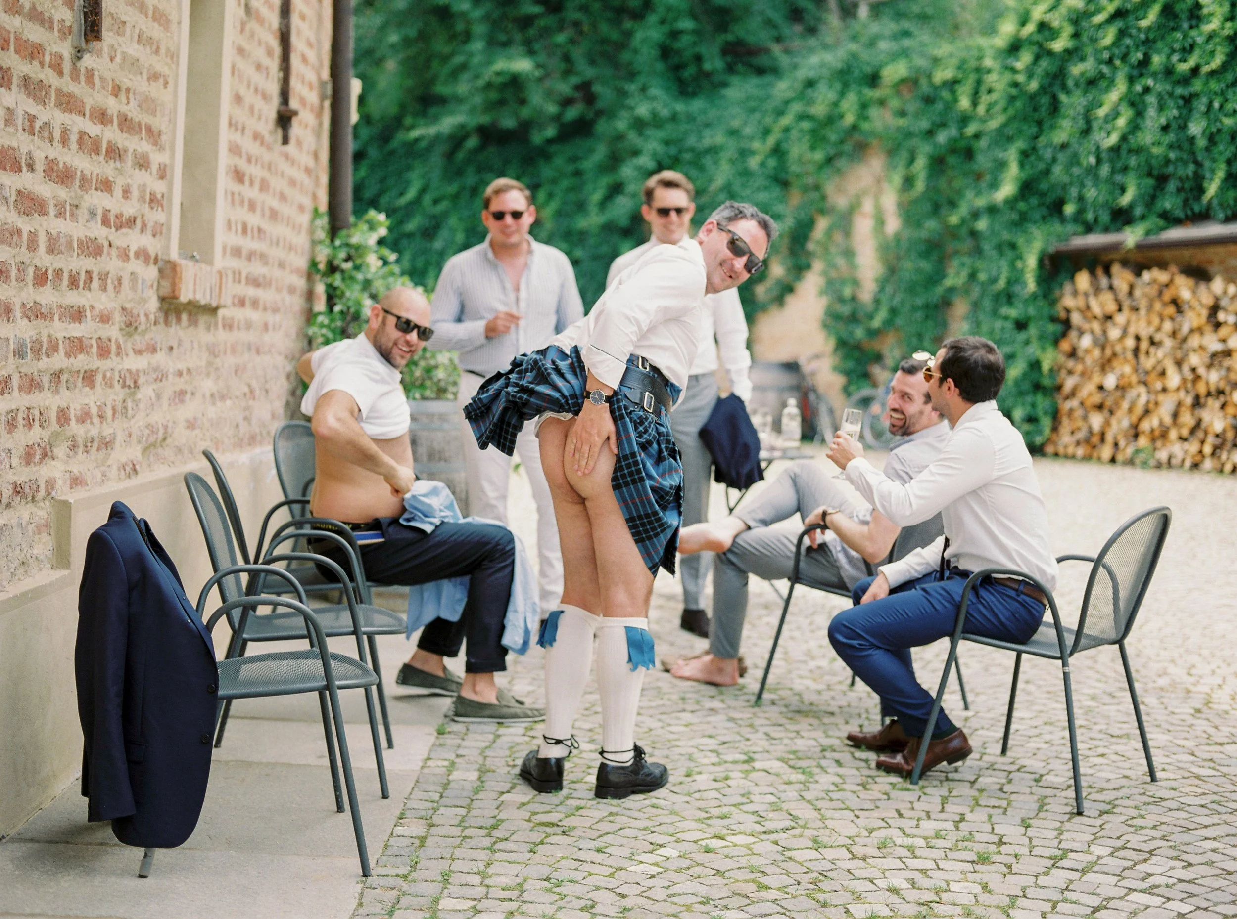 Un gruppo di uomini si diverte a un picnic all'aperto, alcuni sono seduti sulle sedie e altri stanno in piedi, uno di loro indossa una kilt scozzese aperto e sorride mentre gli altri lo guardano o chiacchierano, in un ambiente esterno con muro di mattoni e vegetazione verde in background.