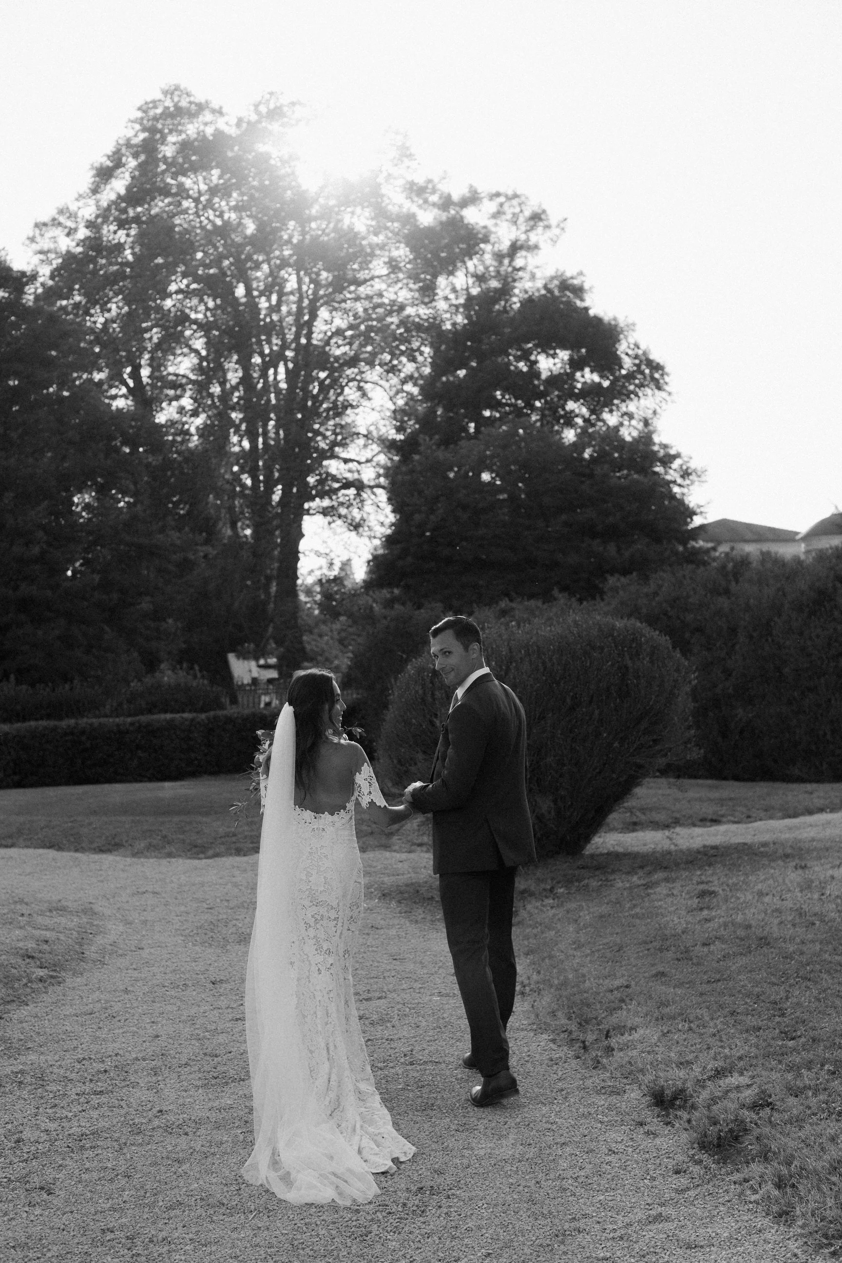 Una coppia in abiti da matrimonio si tiene per mano in un giardino, con grandi alberi e un prato sullo sfondo, sotto un cielo chiaro.