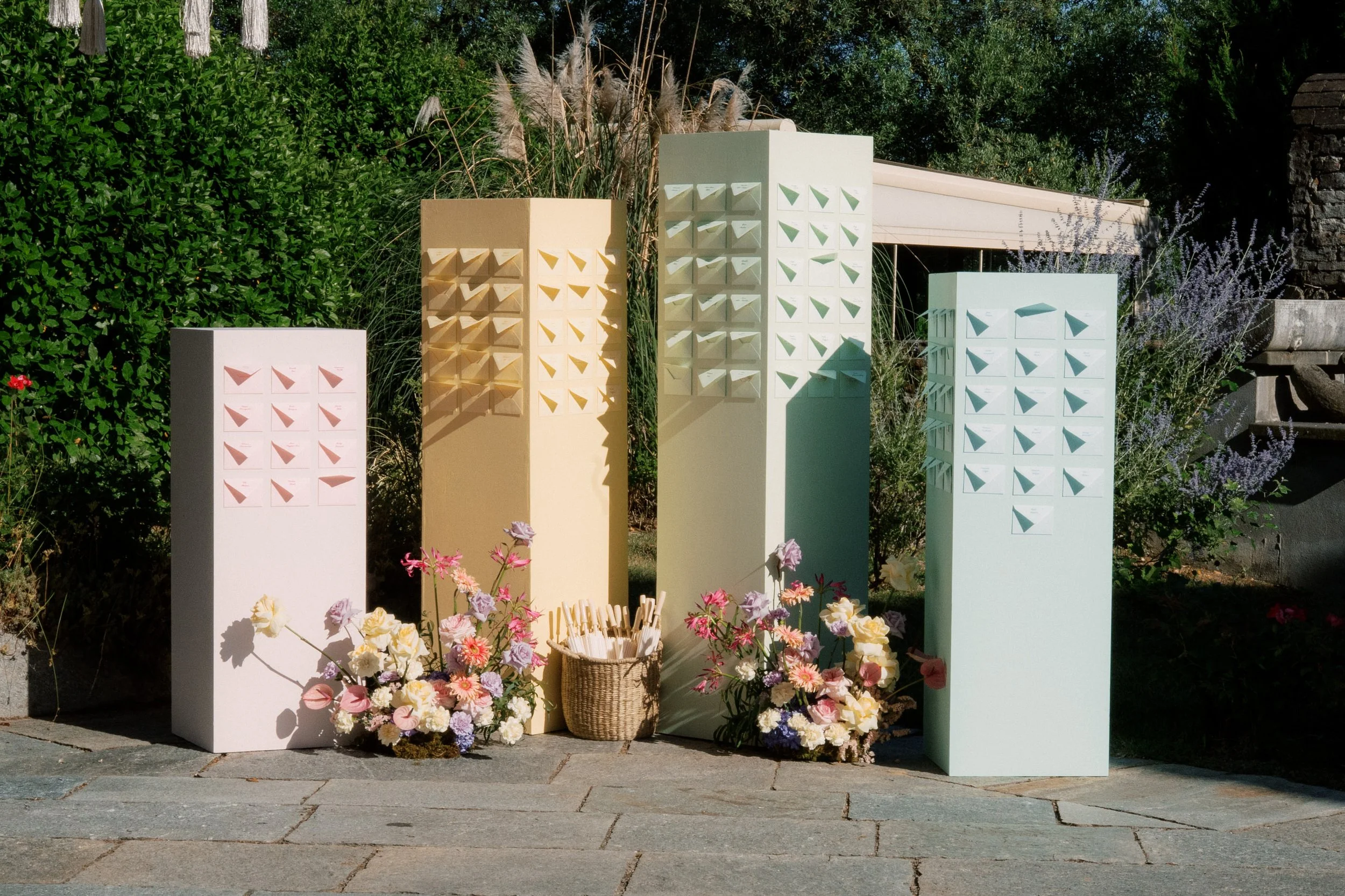 Quattro scaffali di colore bianco, giallo e verde con forme geometriche, decorati con fiori color pastello e un cesto con fiori freschi, in un giardino con vegetazione e panchine in pietra.