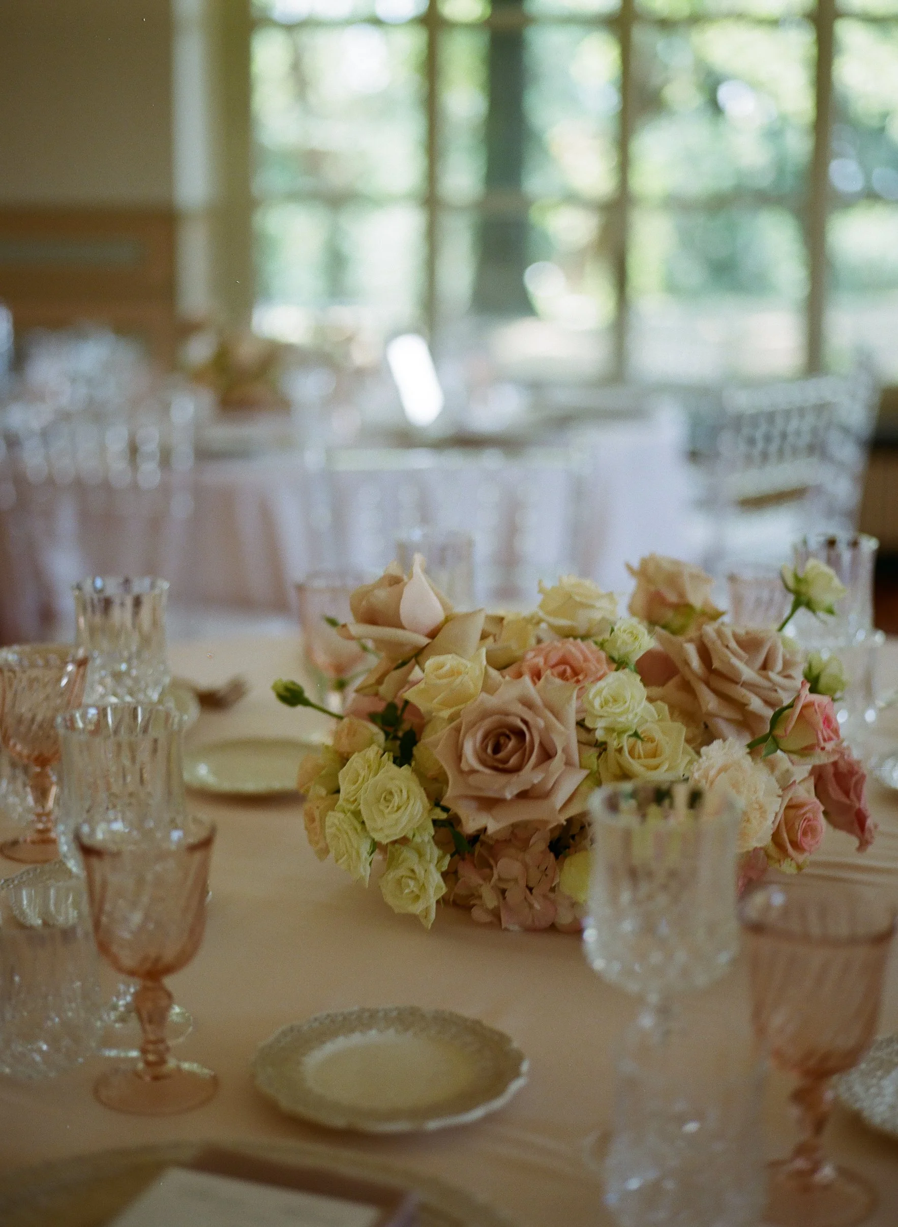 Centro tavola con fiori di rose bianchi, rosa e beige, decorazioni di cristallo rosa e bicchieri, su tavolo per evento o ricevimento, con sfondo di grandi finestre con vista su un giardino verde.