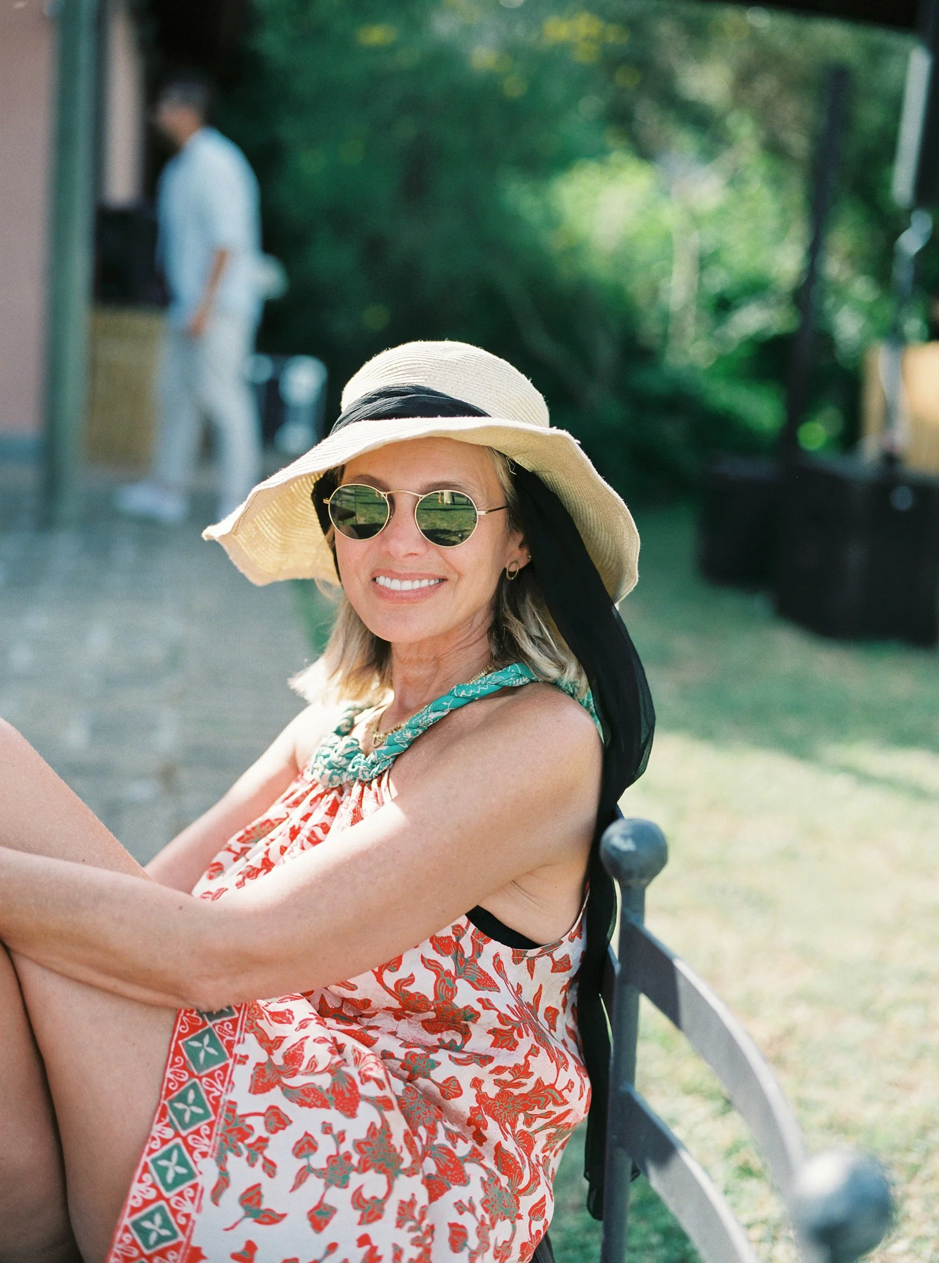 Una donna con cappello di paglia, occhiali da sole, sorriso e vestito estivo a motivo floreale, seduta su una panchina in un parco, in un ambiente estivo soleggiato.