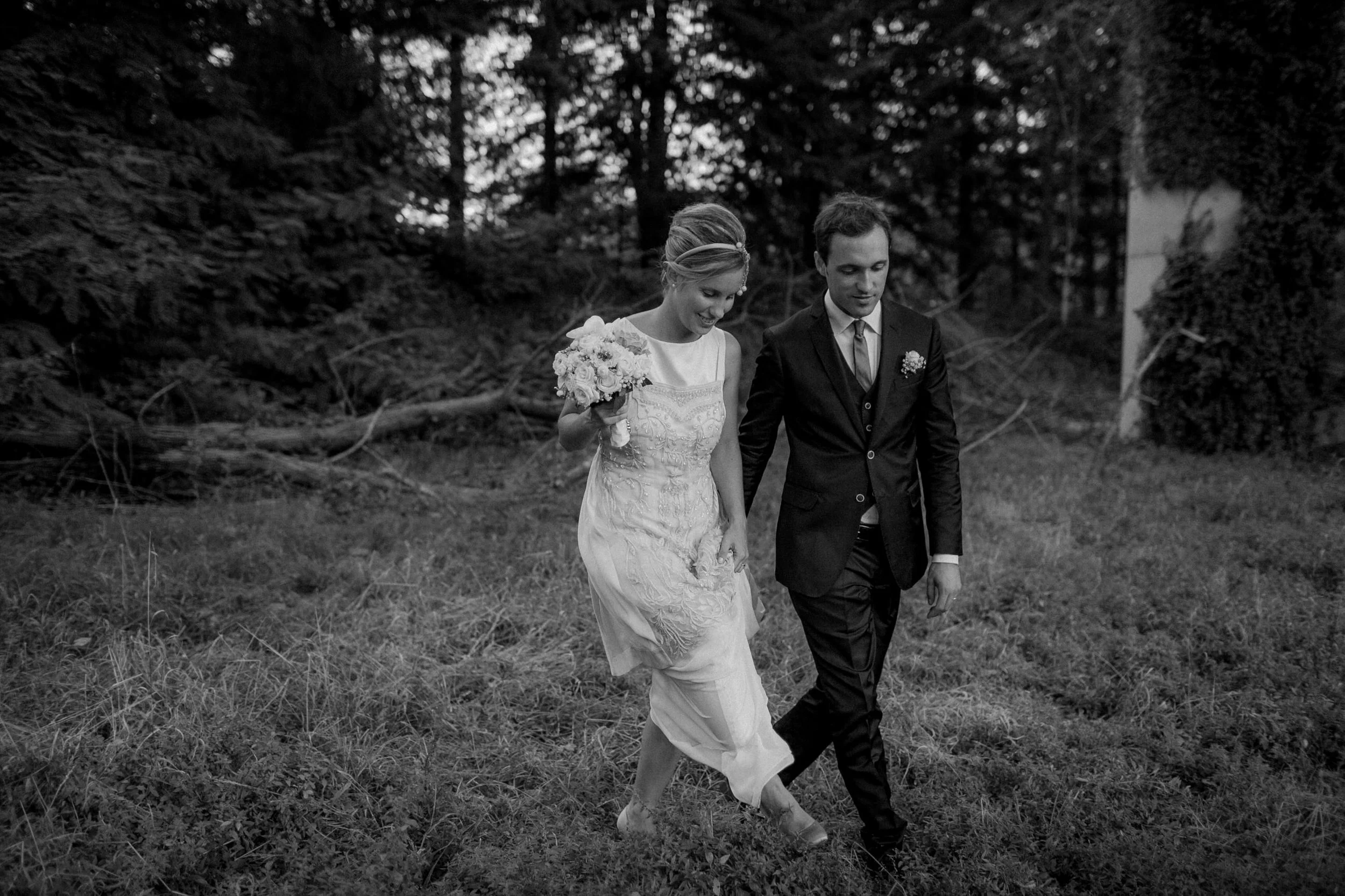 Un uomo e una donna vestiti da sposi camminano insieme in un prato all'aperto, circondati da alberi, in questa foto in bianco e nero.