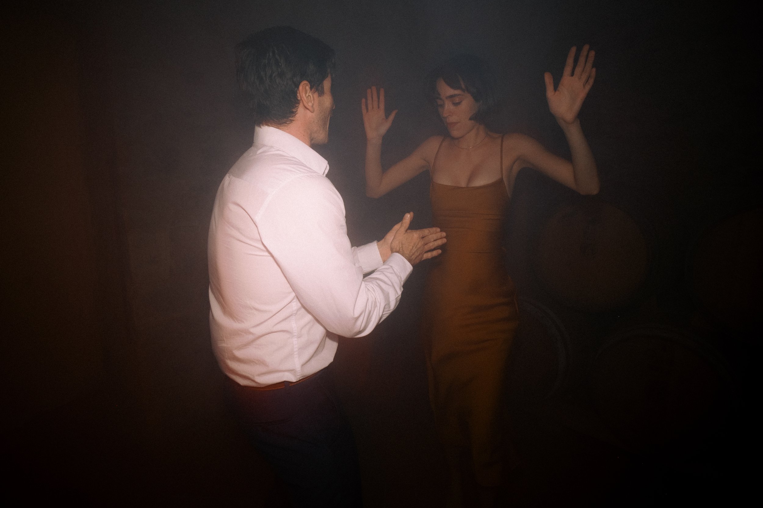Un uomo con camicia bianca e una donna in abito marrone stanno ballando o interagendo in un ambiente oscuro.