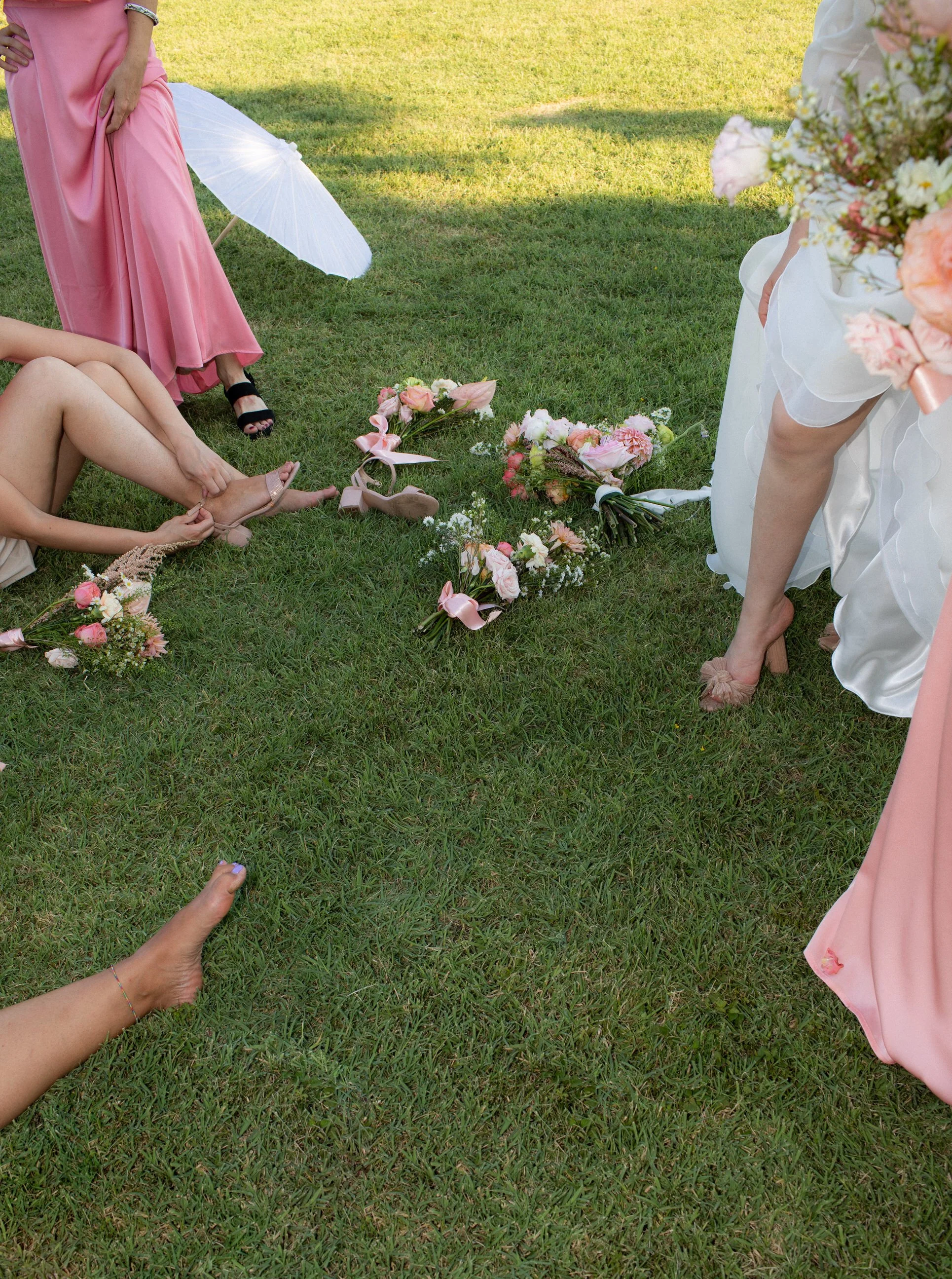 Persone che indossano abiti da cerimonia e scarpe eleganti collaborano per allacciare le scarpe di una donna distesa sull'erba, con fiori e bouquet sul prato.