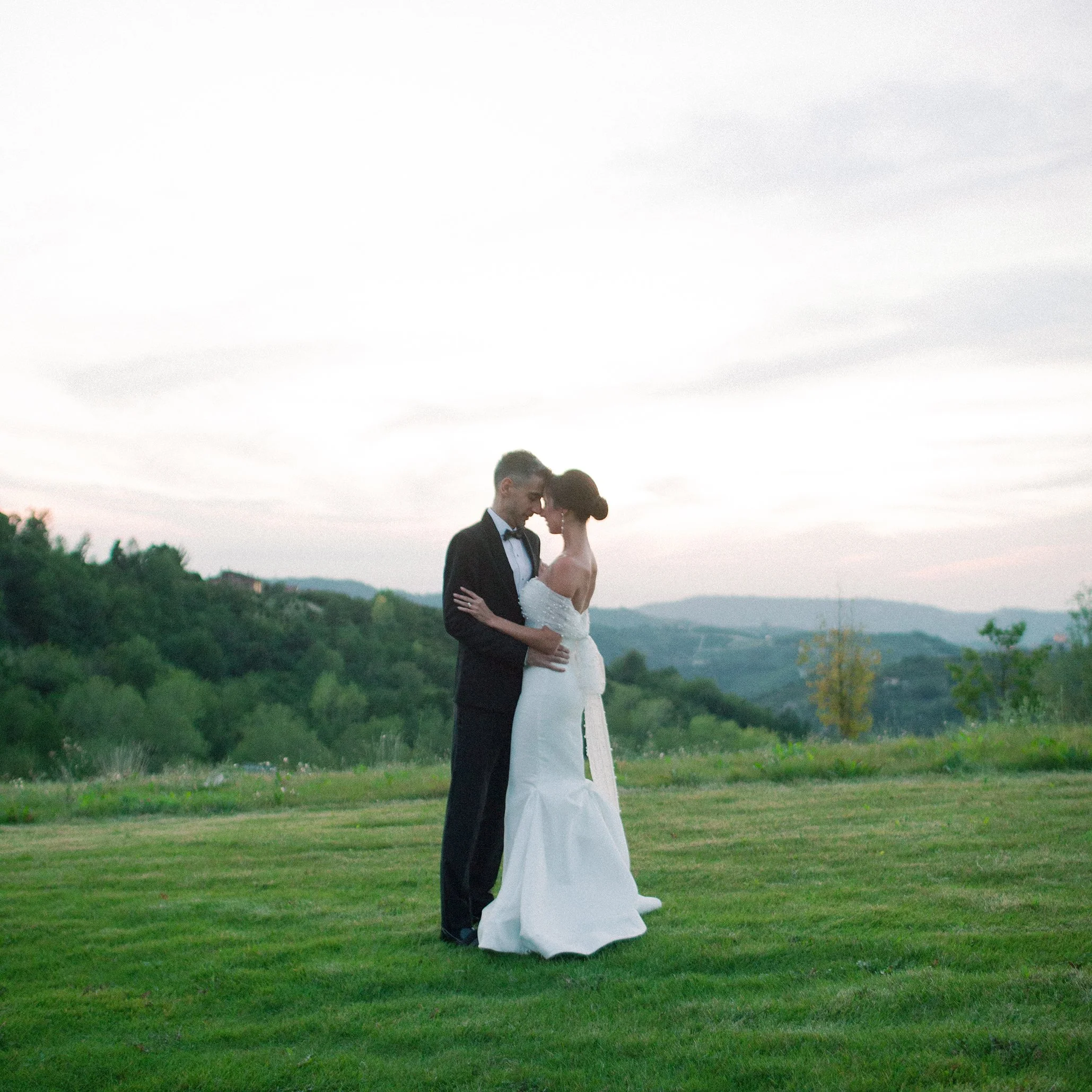 Una coppia di sposi si abbraccia e si bacia in un prato verde, con un cielo nuvoloso e colline sullo sfondo, durante un matrimonio all'aperto.