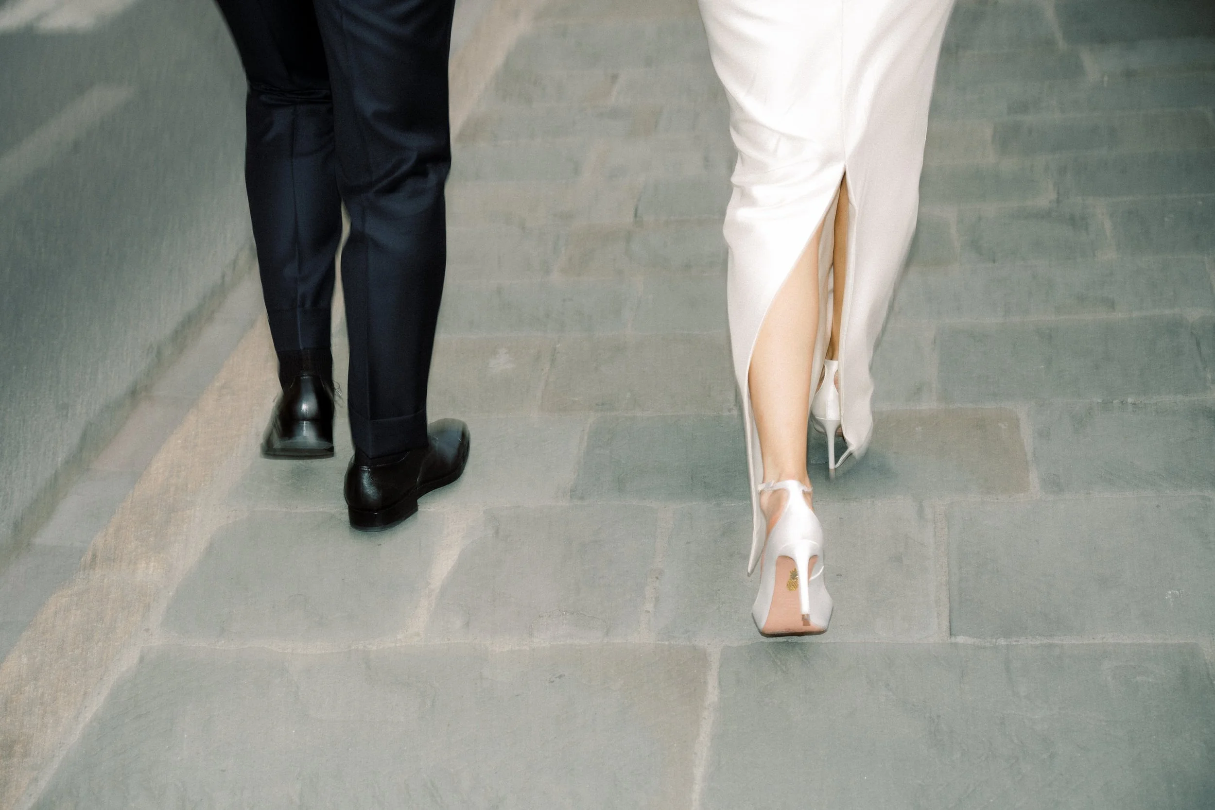 Parte inferiore di due persone che camminano sul pavimento in pietra, una vestita con pantaloni neri e scarpe nere, l'altra con un vestito bianco lungo e scarpe con tacco alto e cinturino alla caviglia.