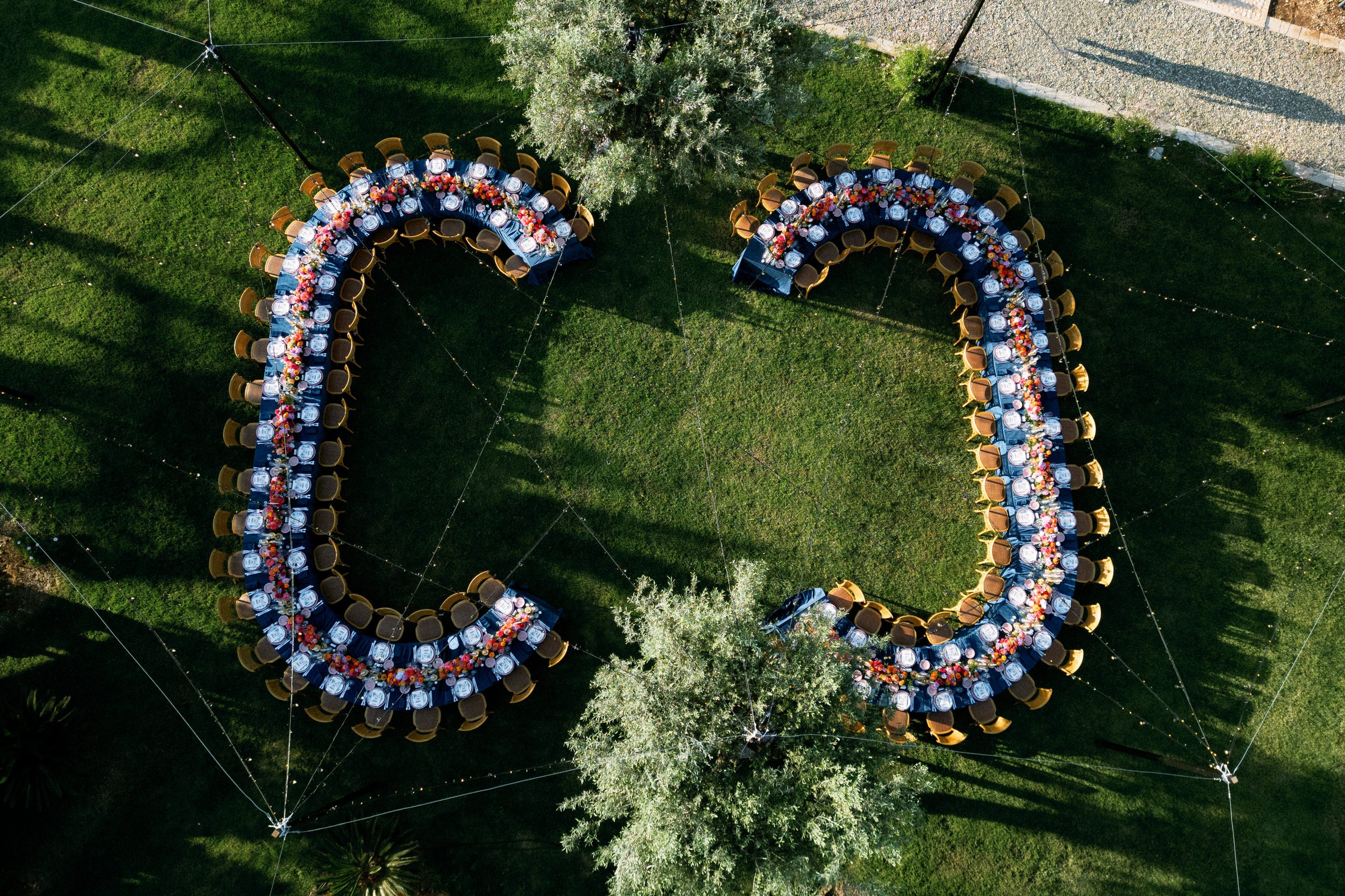 Tavola apparecchiata in un giardino decorato con fiori colorati, con sedie di legno, set di piatti e bicchieri, in un evento all'aperto.