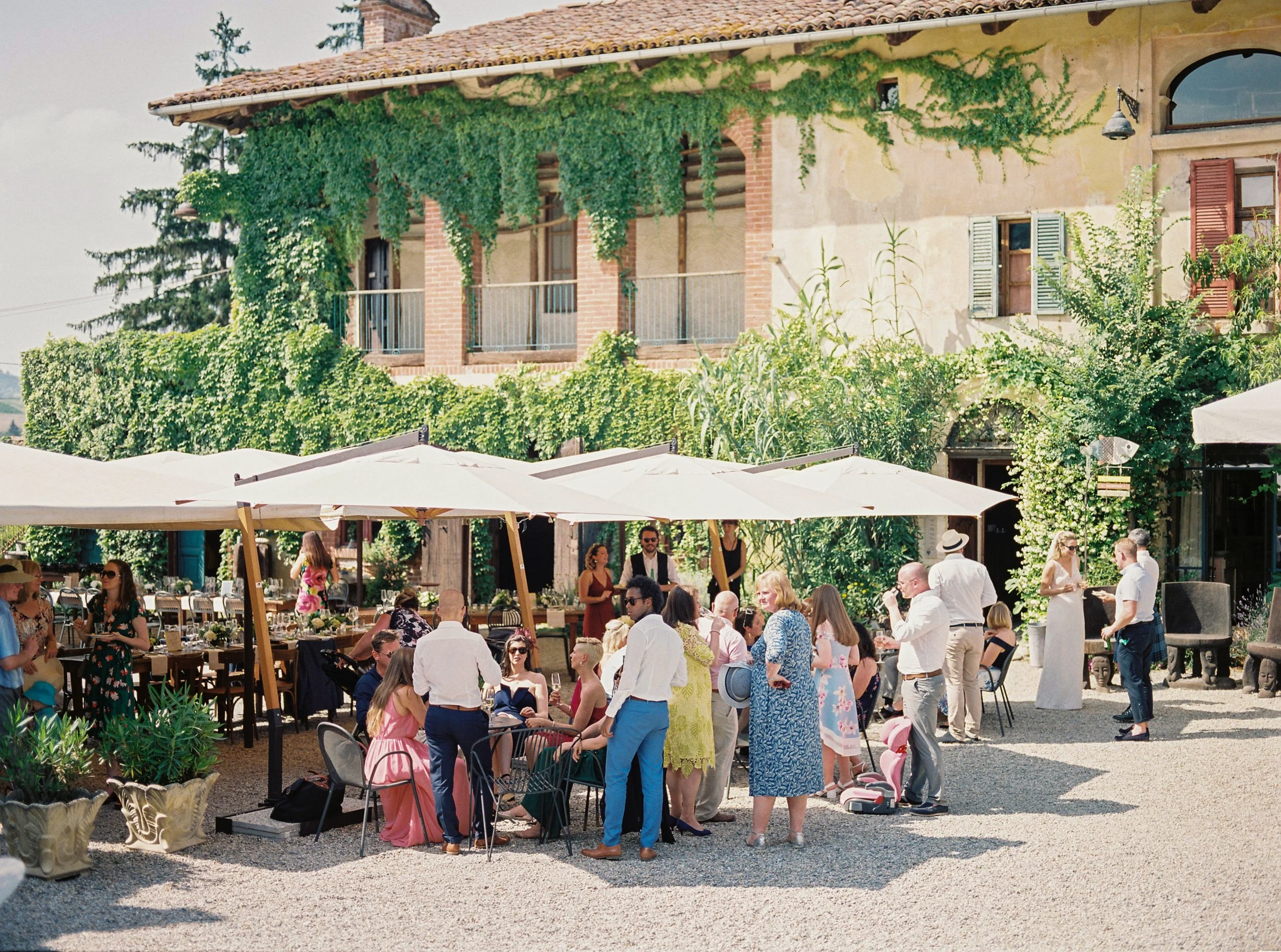 Festa all'aperto con molte persone riunite, alcune sedute a tavoli e altre in piedi, sotto grandi ombrelloni in un cortile con un edificio storico in pietra e piante verdi lussureggianti sullo sfondo.