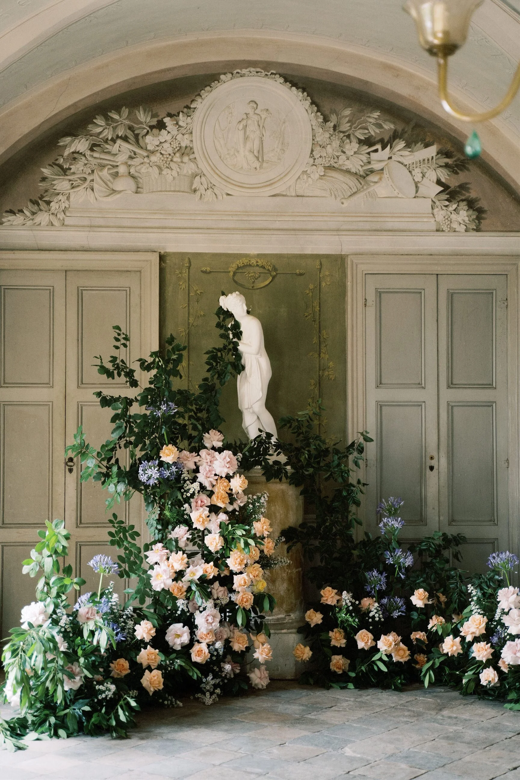 Un'area decorata con molte piante e fiori, con una statua bianca di una donna con un abito lungo, riparata da un architrave con decorazioni e un fregio raffigurante una scena classica e motivi di fiori e frutti.