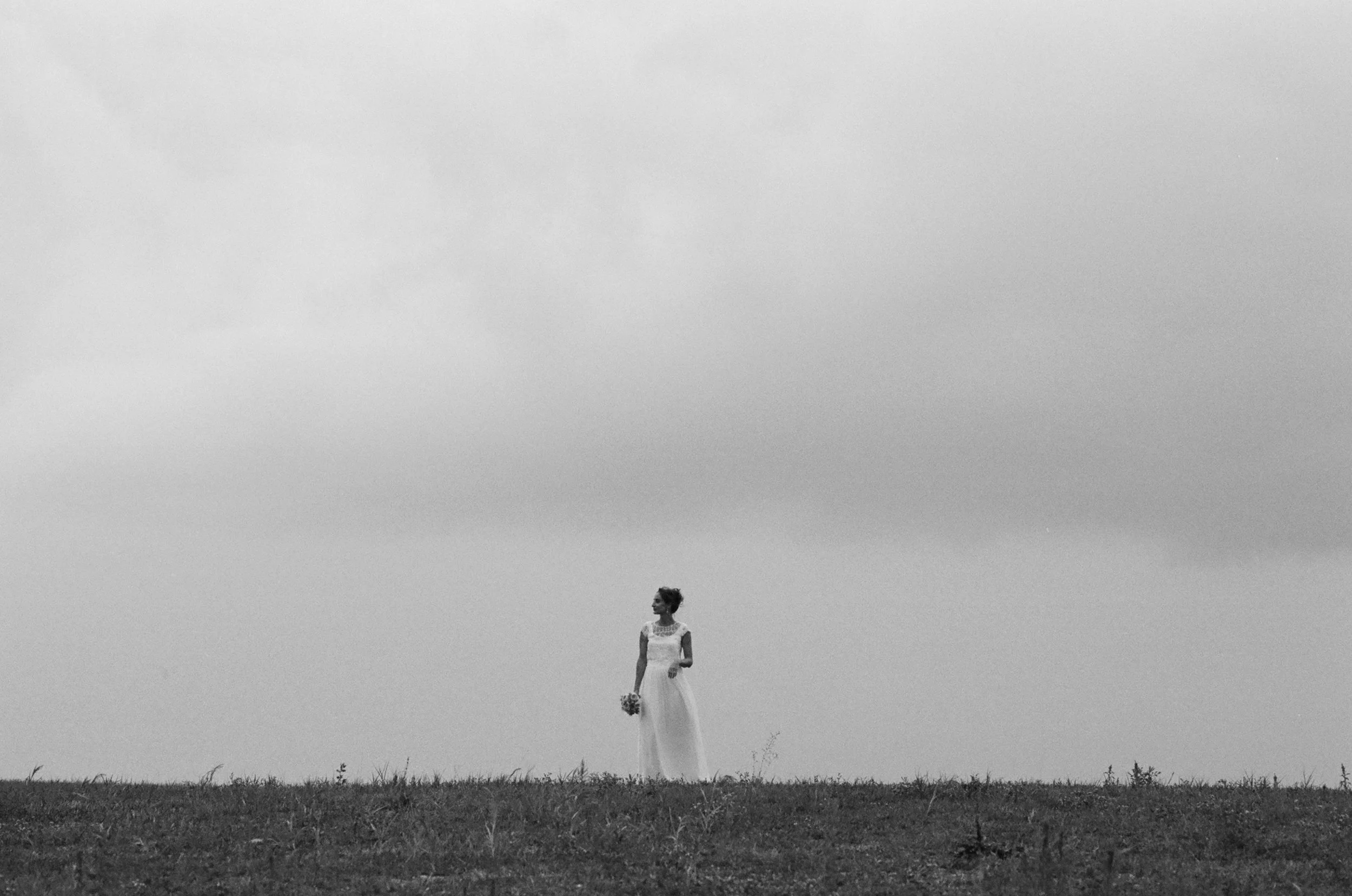 Una donna in abito bianco che tiene un mazzo di fiori, si trova in un campo con l'erba, sotto un cielo grigio, in un contesto in bianco e nero.