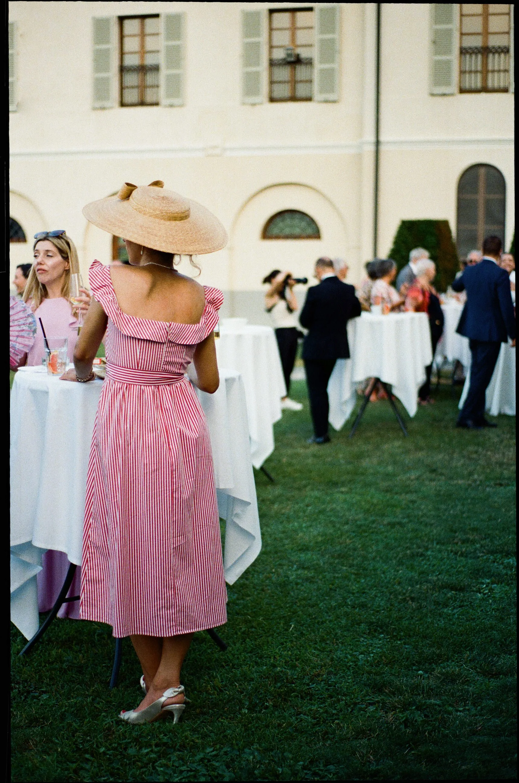 Donne che socializzano a un evento all'aperto, una donna indossa un vestito rosa a strisce e un cappello di paglia, mentre altre persone sono al di sotto di un gazebo, sul prato verde.