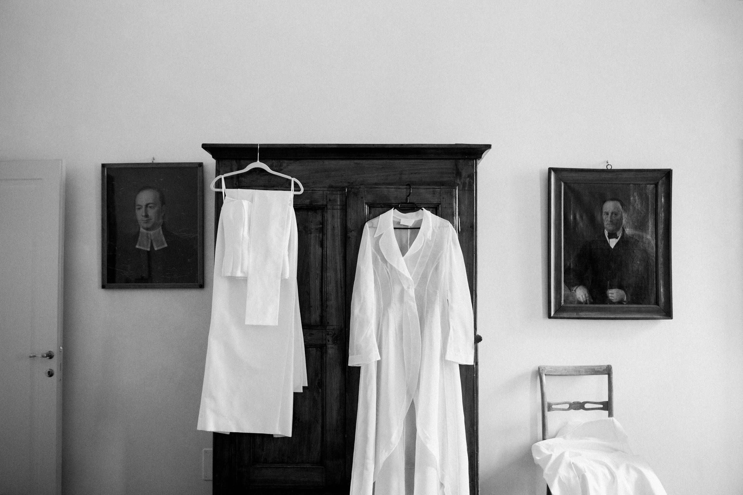 Abbigliamento bianco appeso su un armadio e un letto con lenzuolo, con ritratti in cornice alle pareti.