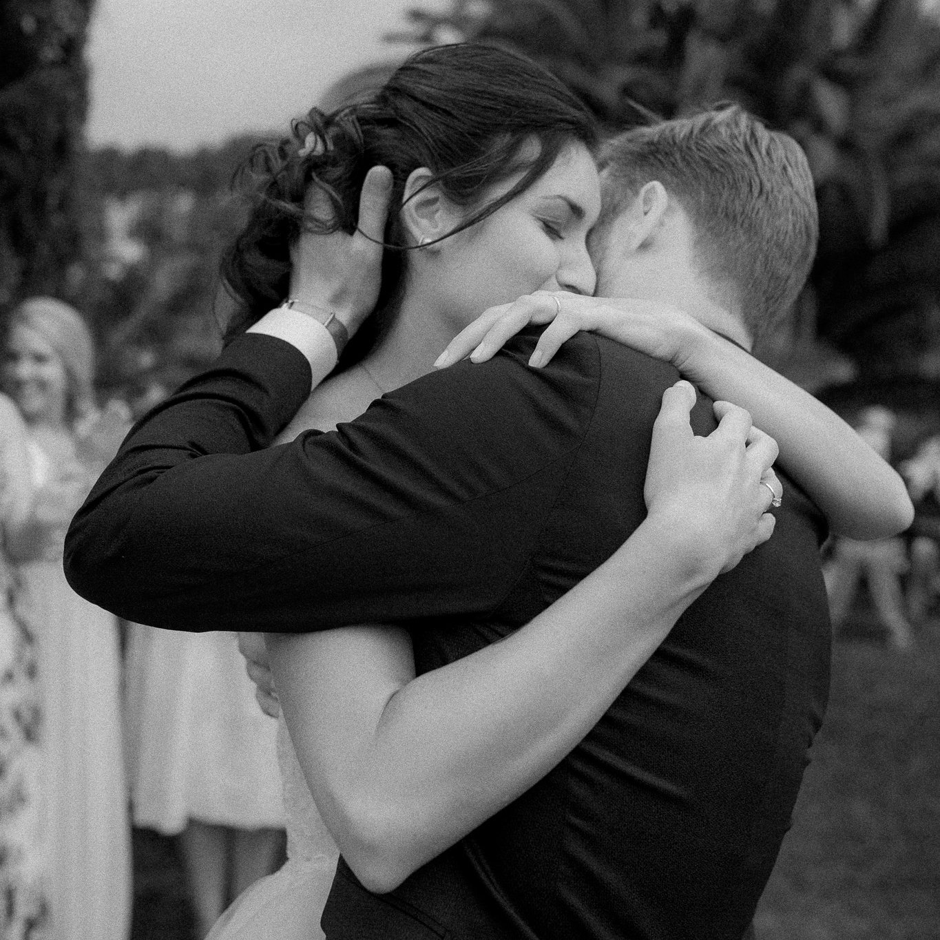 Due persone che si abbracciano, probabilmente in un momento di festa o matrimonio, in un ambiente all'aperto con persone sullo sfondo.