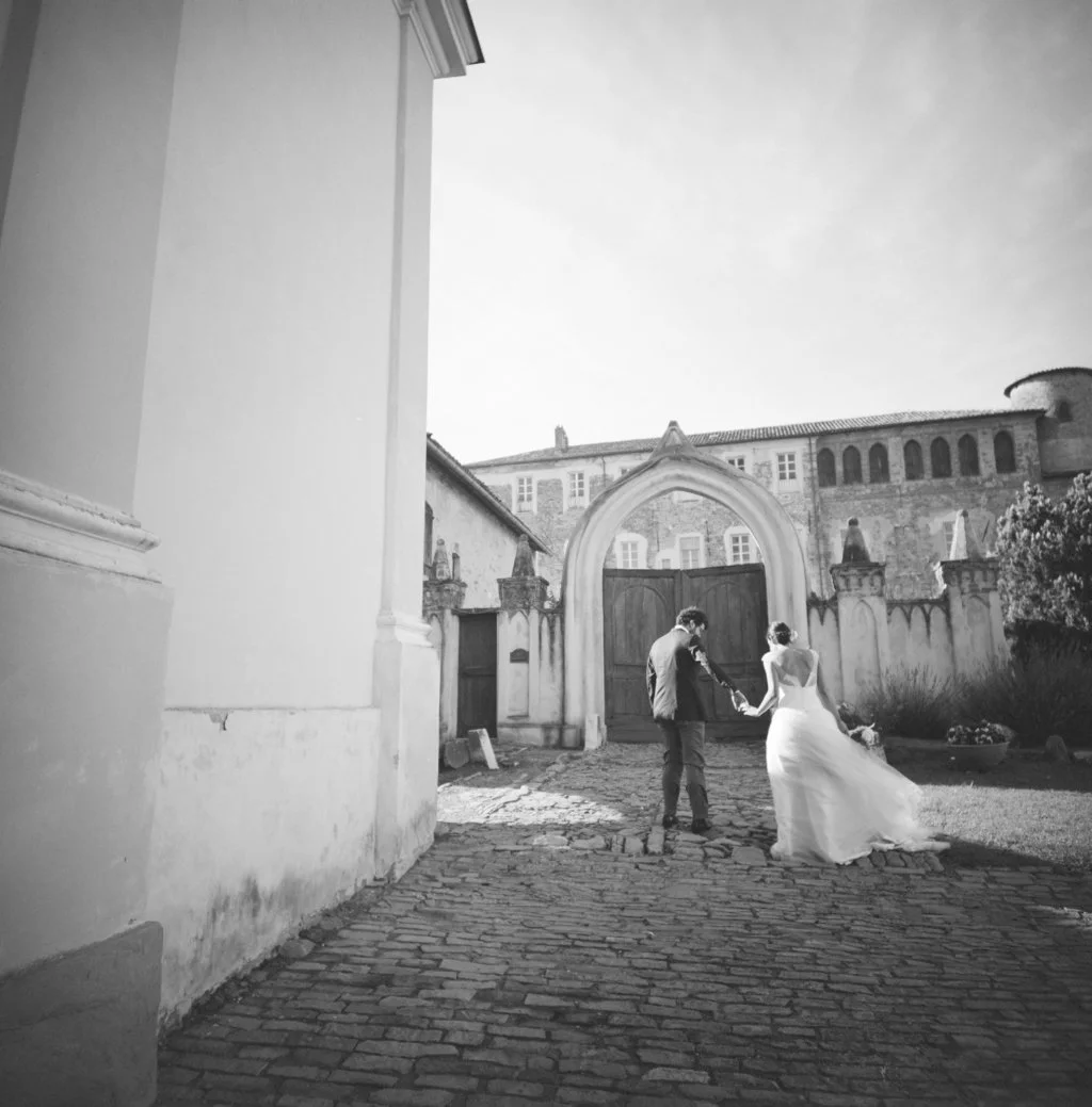 Una coppia di sposi si tiene per mano davanti a un grande cancello in un cortile storico, in un'immagine in bianco e nero.