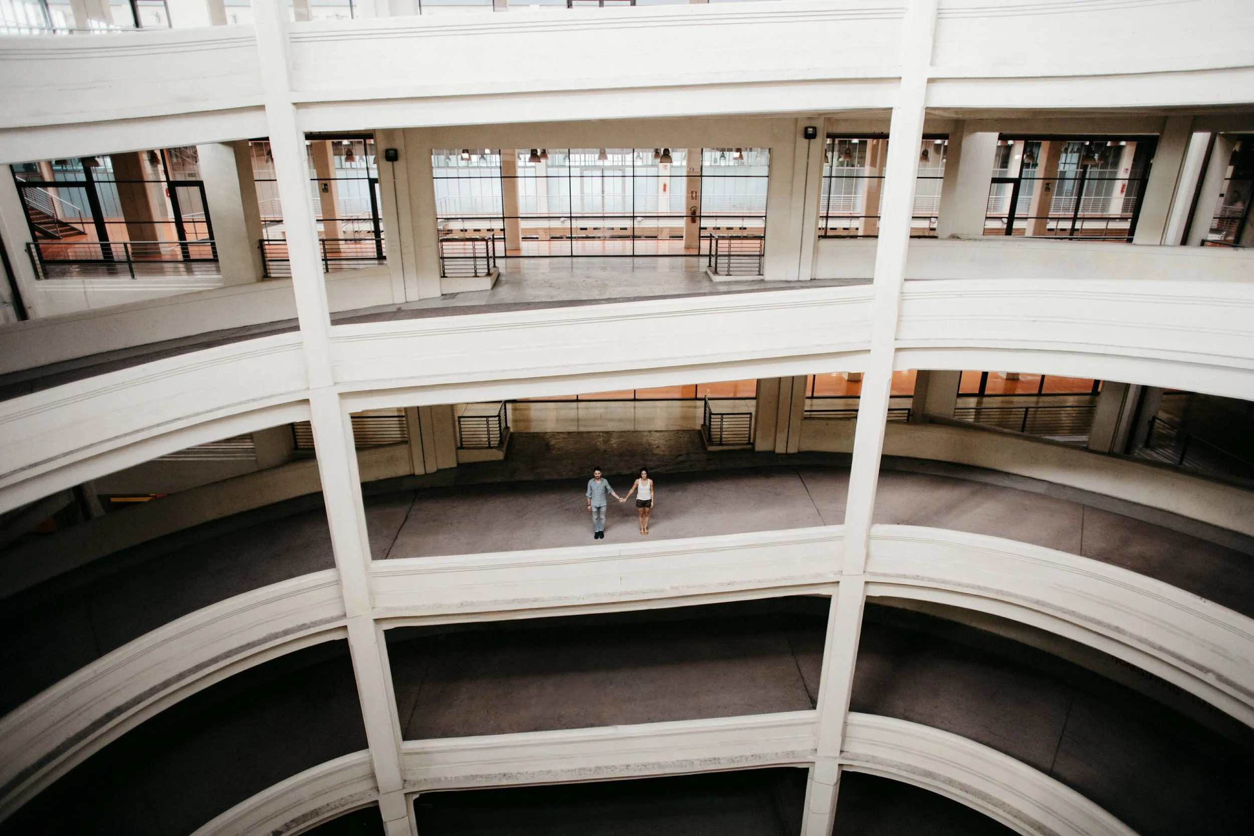 Due persone tengono per mano nel cortile di un edificio moderno con tre piani visibili, tutti con balconi curvi e ringhiere di metallo, su un'architettura bianca.