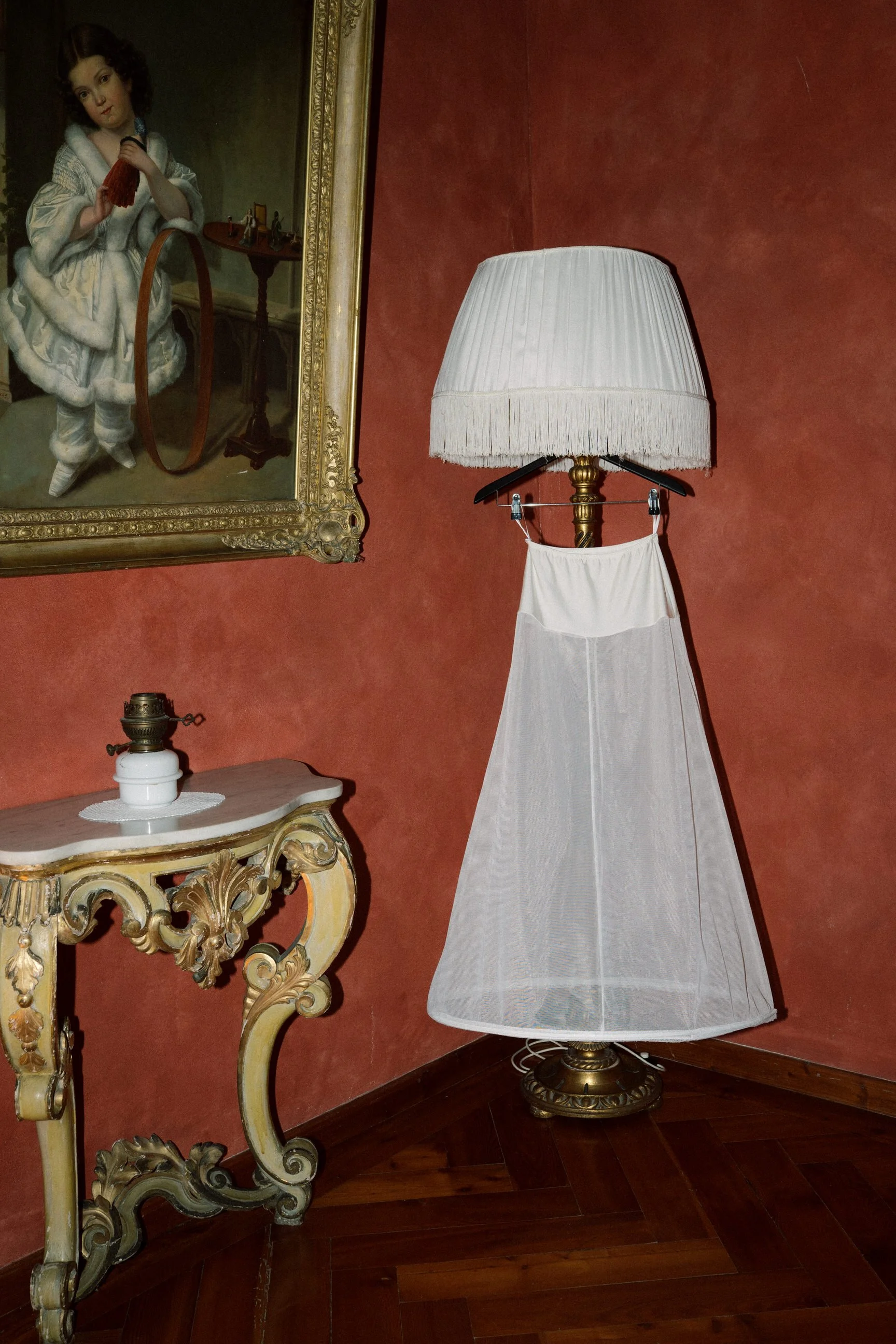 Una stanza con una lampada con base dorata e paralume bianco, un quadro con cornice dorata raffigurante una ragazza in un abito d'epoca, una consolle dorata con dettagli artistici e un oggetto decorativo, e un pavimento in legno.